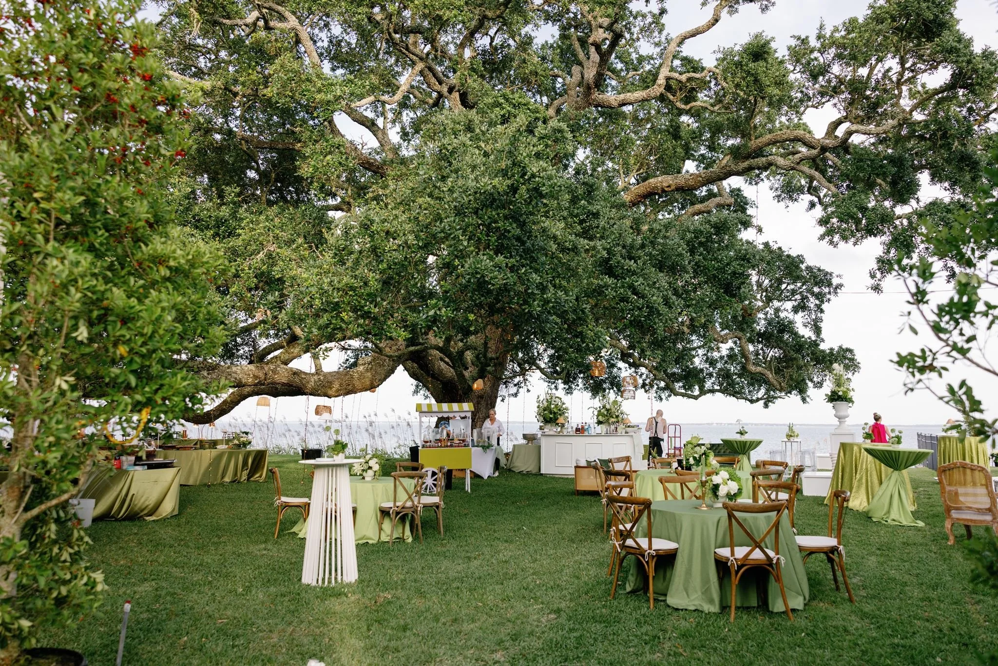 An outdoor event setup with green tables and chairs under a large tree near a waterfront. There are people preparing food behind a white bar, with some in the background. The scene is decorated with flowers and the sky appears overcast.