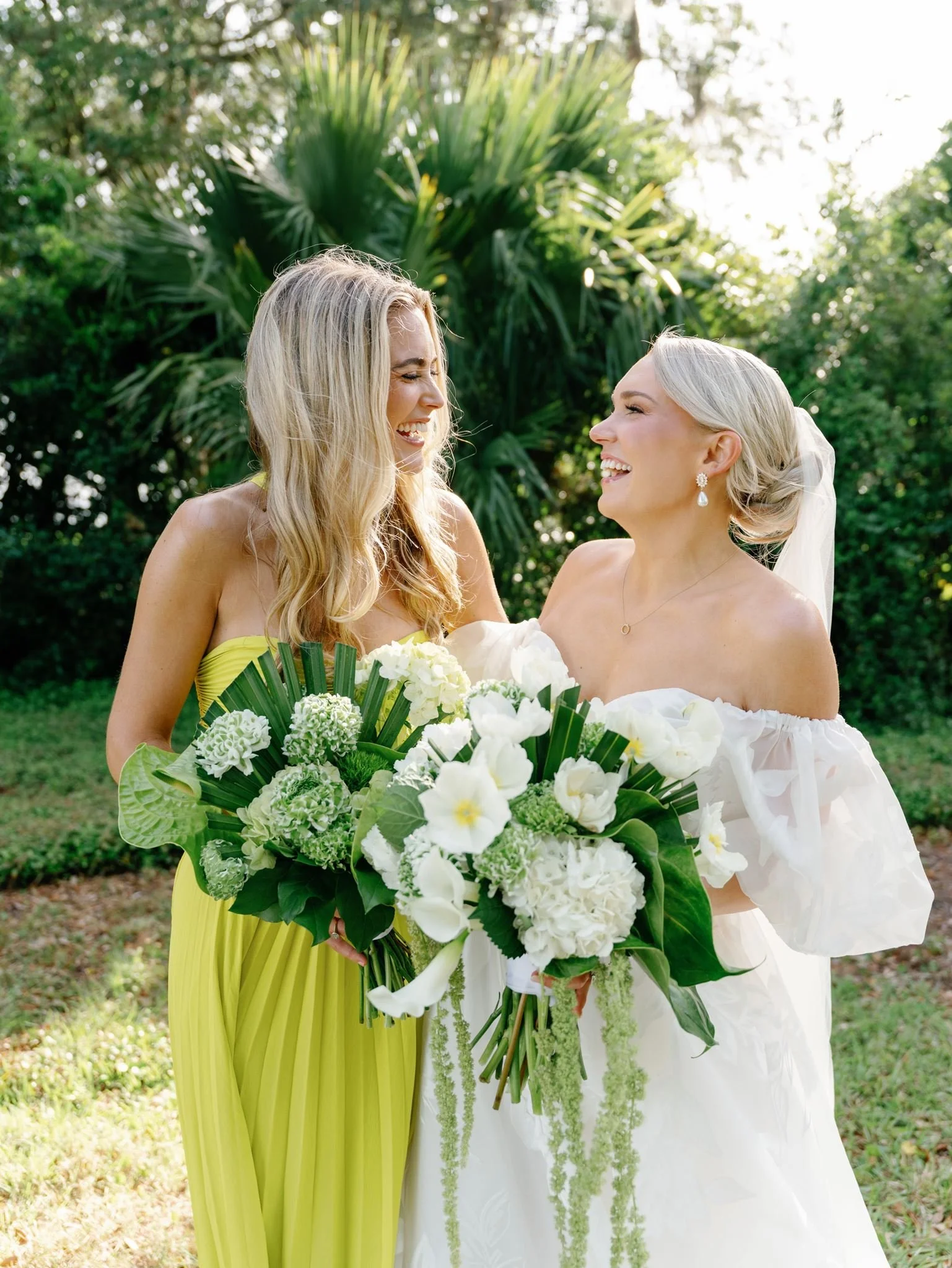 Two women in wedding dresses smiling and holding bouquets of white flowers, standing outdoors with lush green foliage in the background.