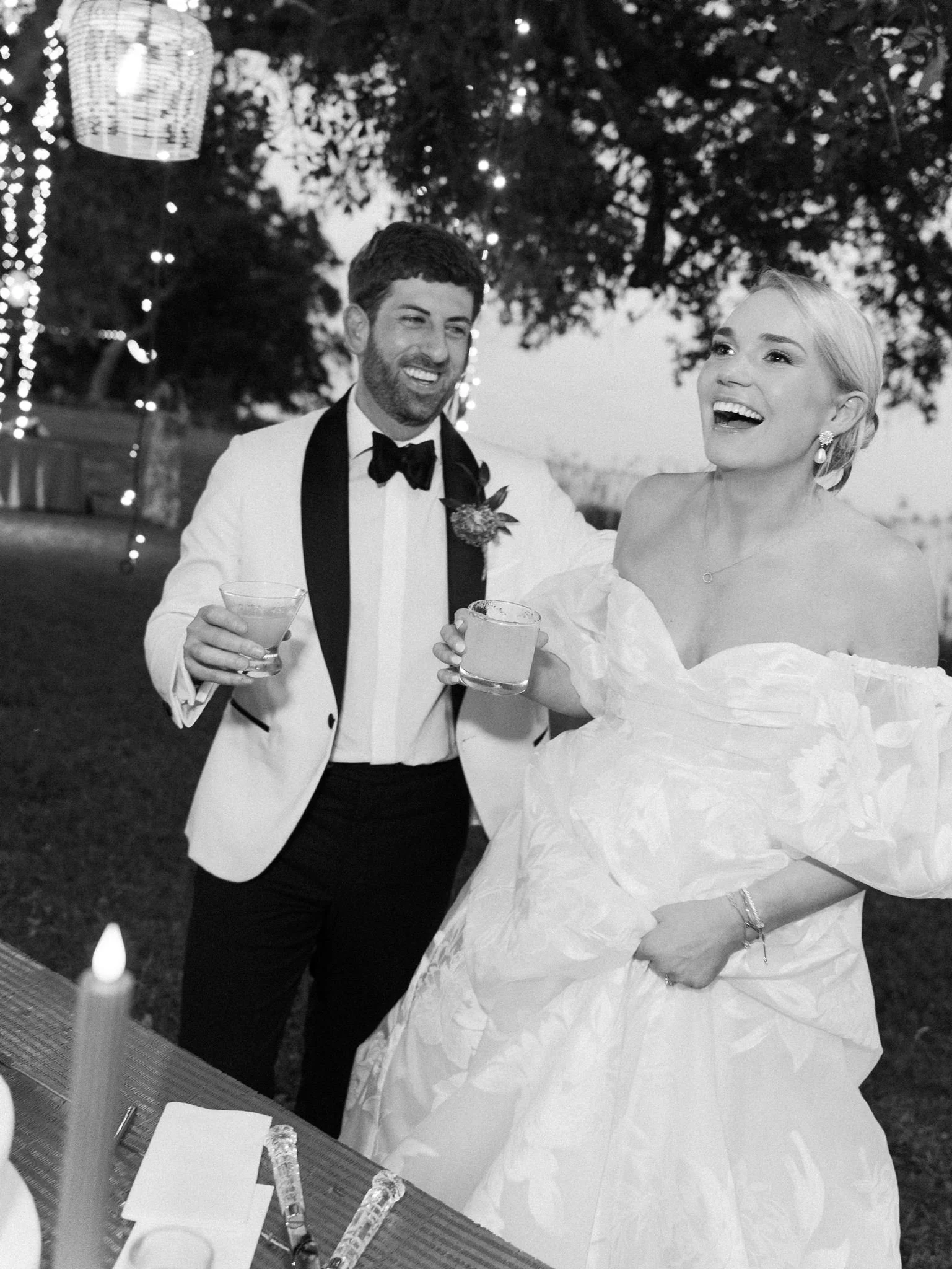 A man and woman dressed in formal attire are celebrating together outdoors, holding drinks and smiling at each other at night with decorative lights in the background.