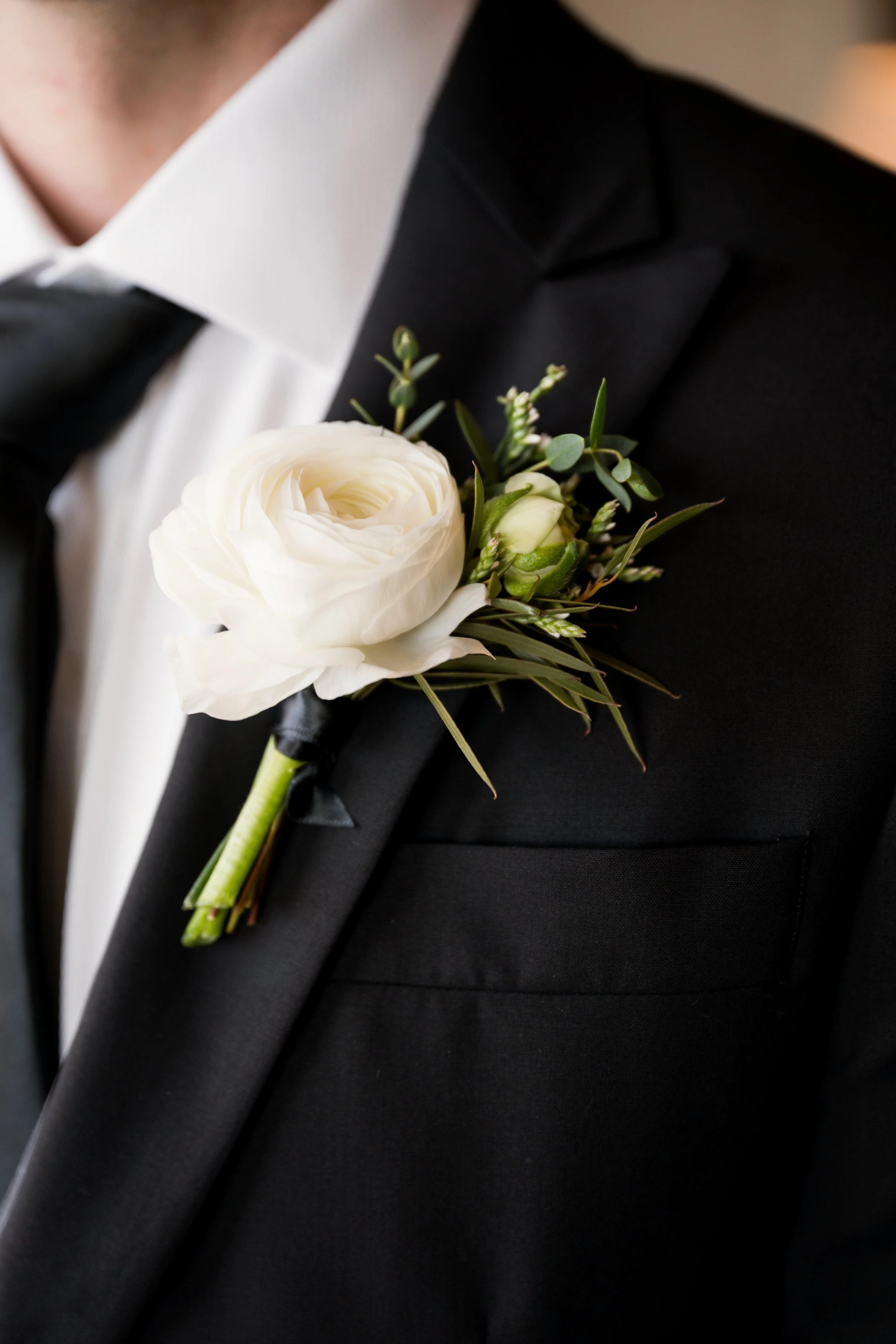 Close-up of a groom in a black tuxedo, white shirt, and black bow tie, wearing a white floral boutonniere on his lapel.