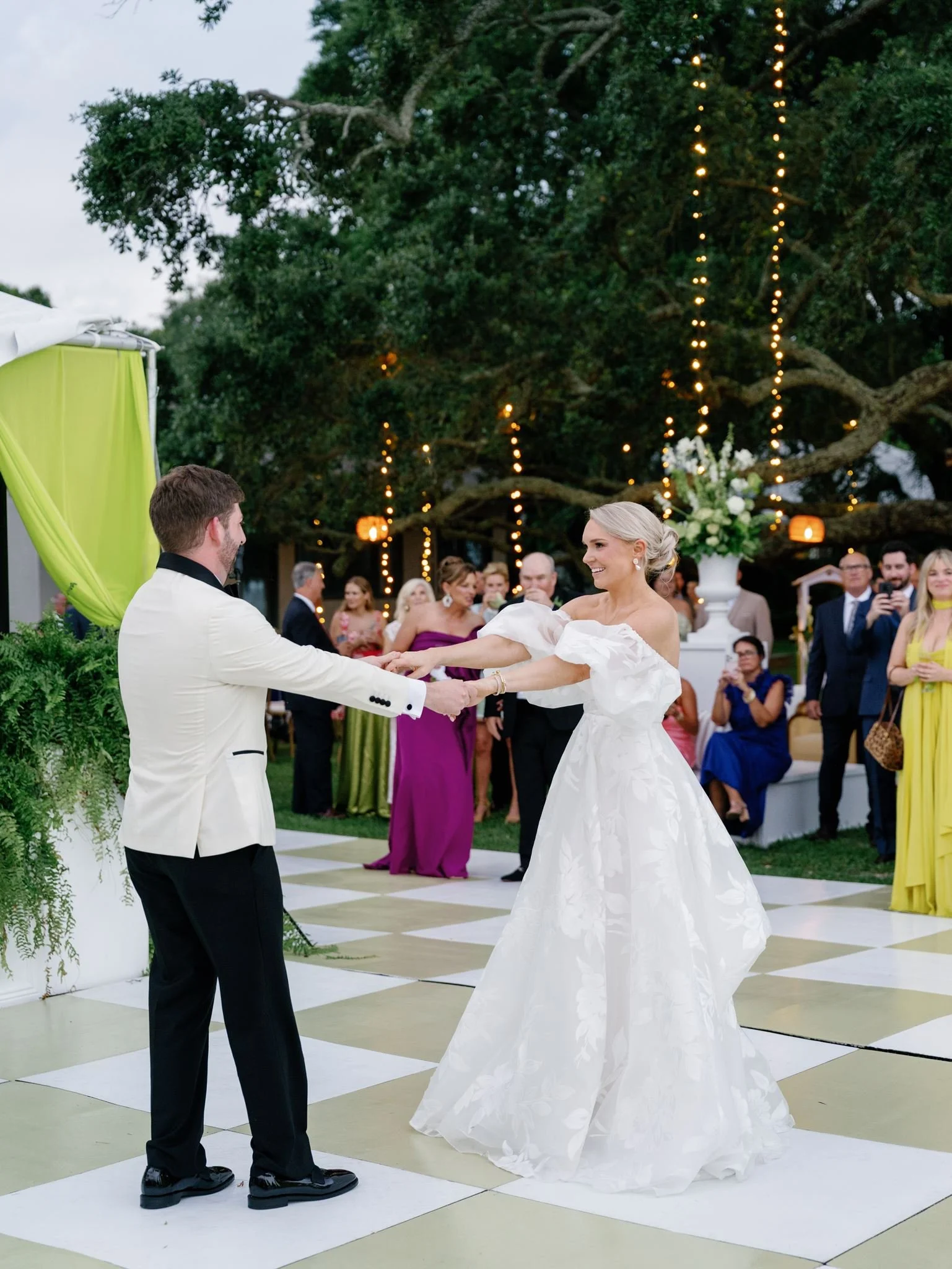 A wedding ceremony taking place outdoors with the bride and groom holding hands and smiling at each other, surrounded by guests and decorated with string lights and floral arrangements.