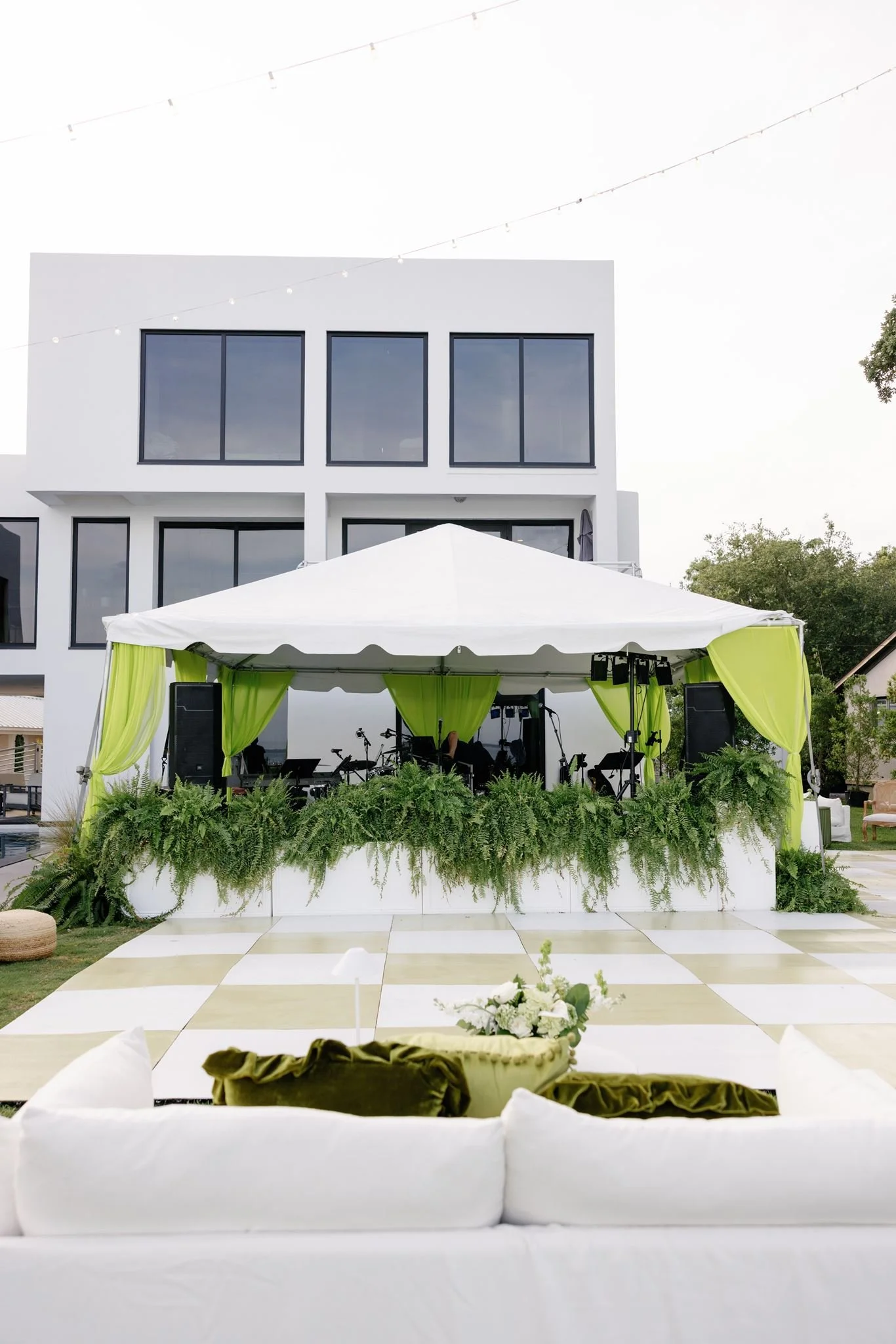 Outdoor event setup with a white stage draped with green curtains, surrounded by lush greenery, with a modern white building with large windows in the background.