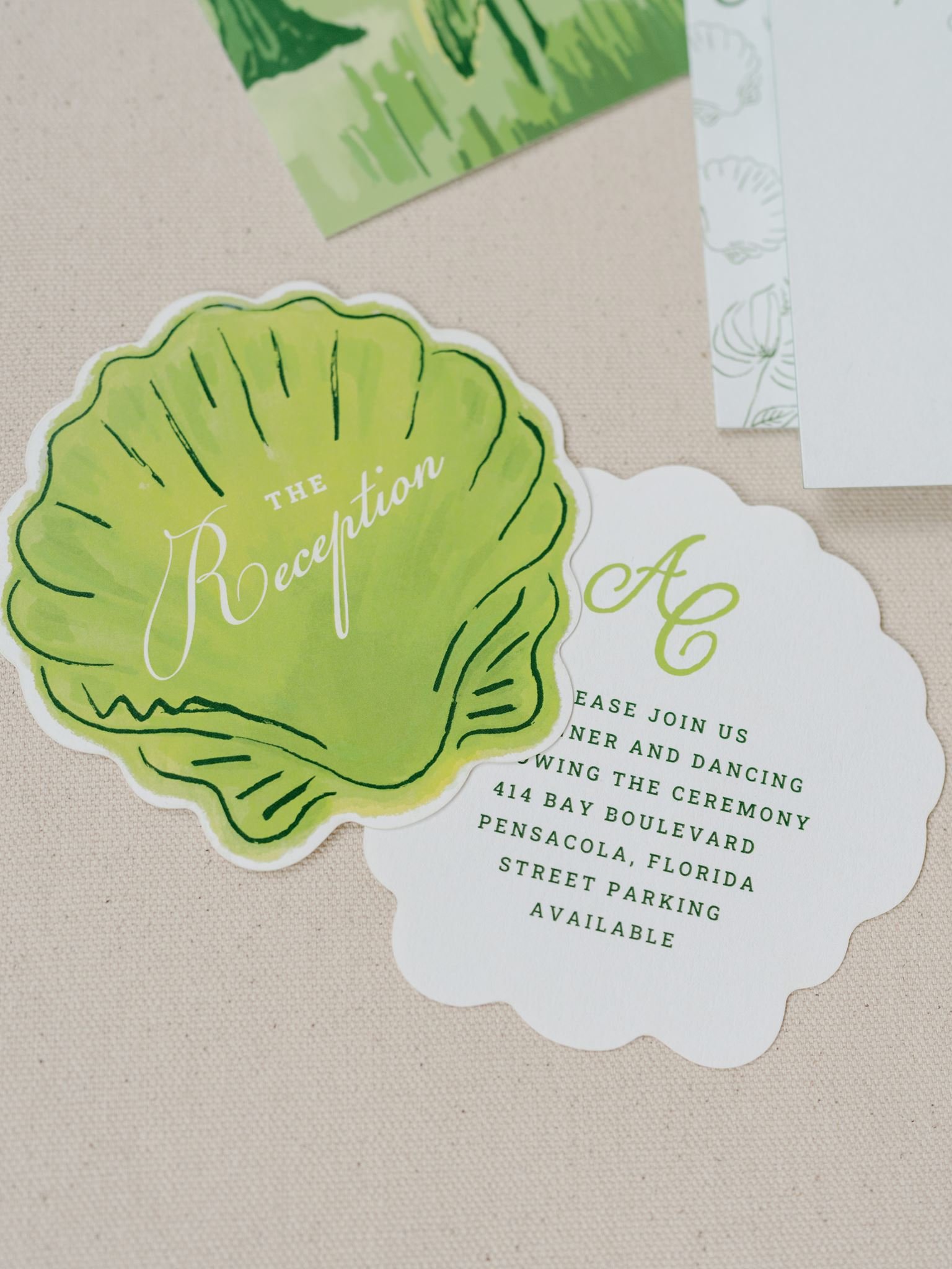 A green clam-shaped sticker with the text 'The Reception' on it, placed on a beige surface, along with a white card with event details and part of a printed paper in the background.