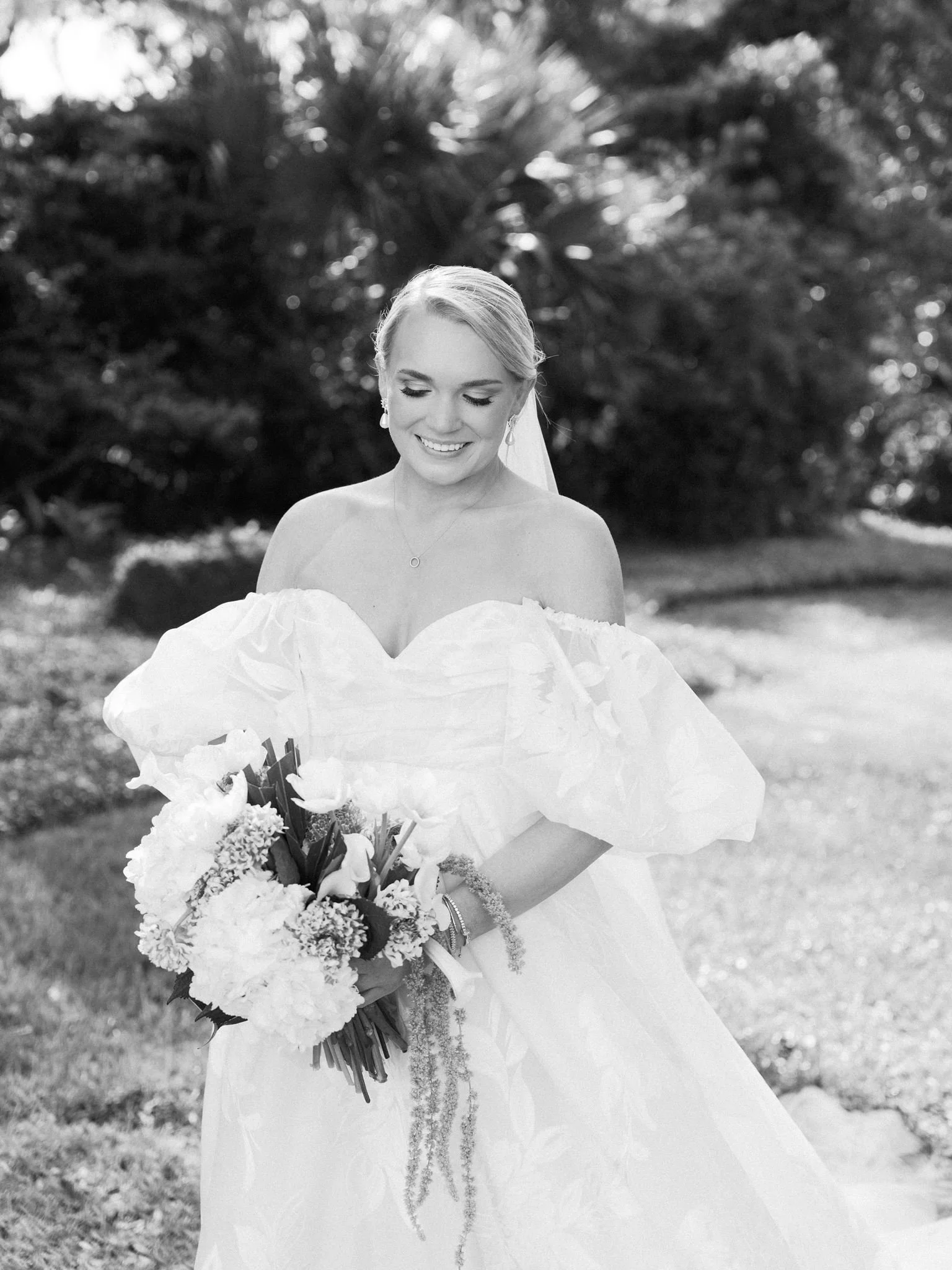 A smiling bride in a strapless wedding gown holding a bouquet of flowers outdoors with trees and natural surroundings in the background.