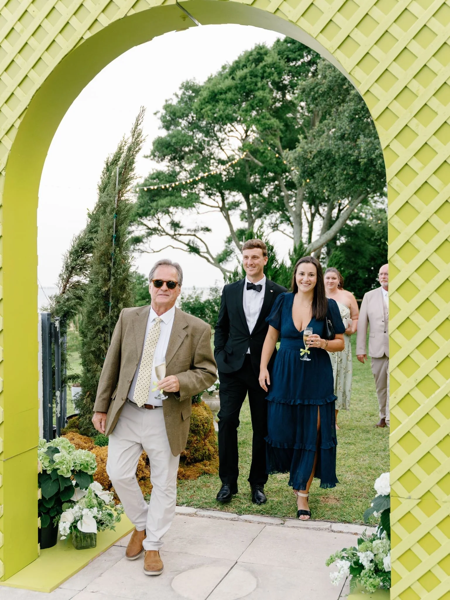 People walking through a decorated outdoor archway at a wedding or event, with greenery and trees in the background.