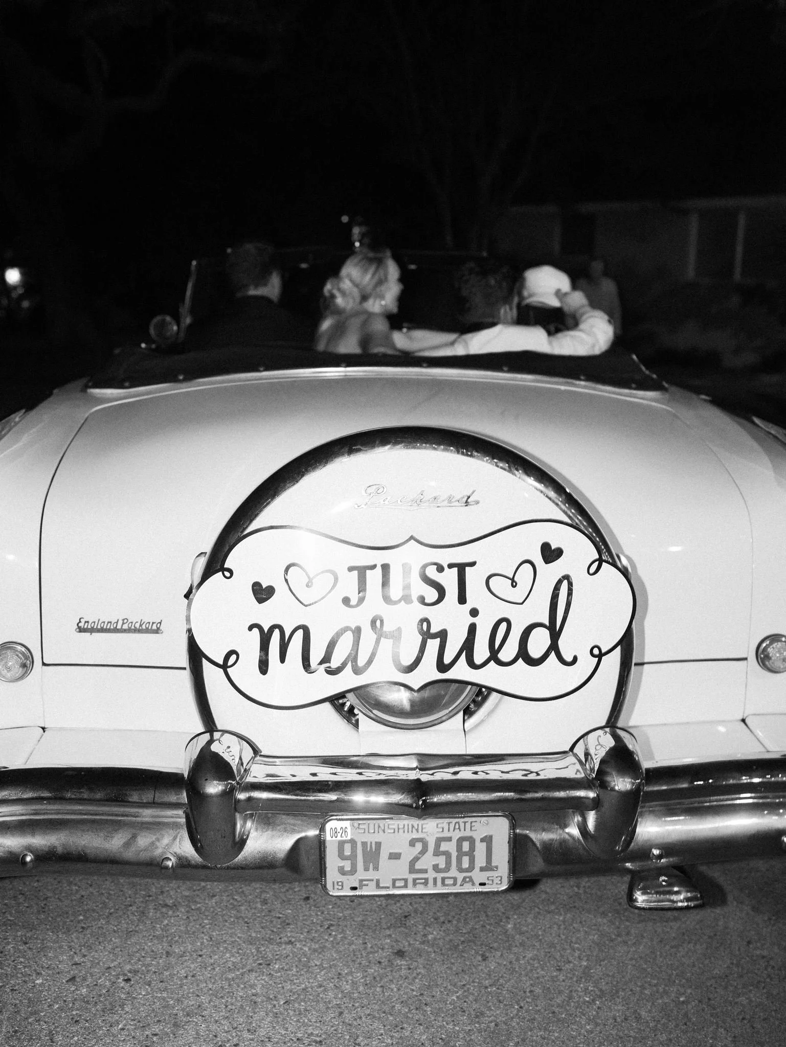 Back of a vintage car with a large 'Just Married' sign on the trunk, a Florida license plate reading '9W-2581,' and several people sitting in the car during nighttime.