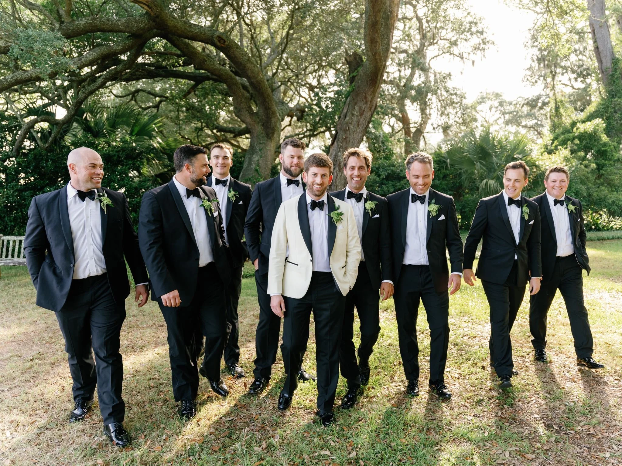 A group of men dressed in formal black tuxedos walking outdoors in a park-like setting with large trees and green foliage in the background.