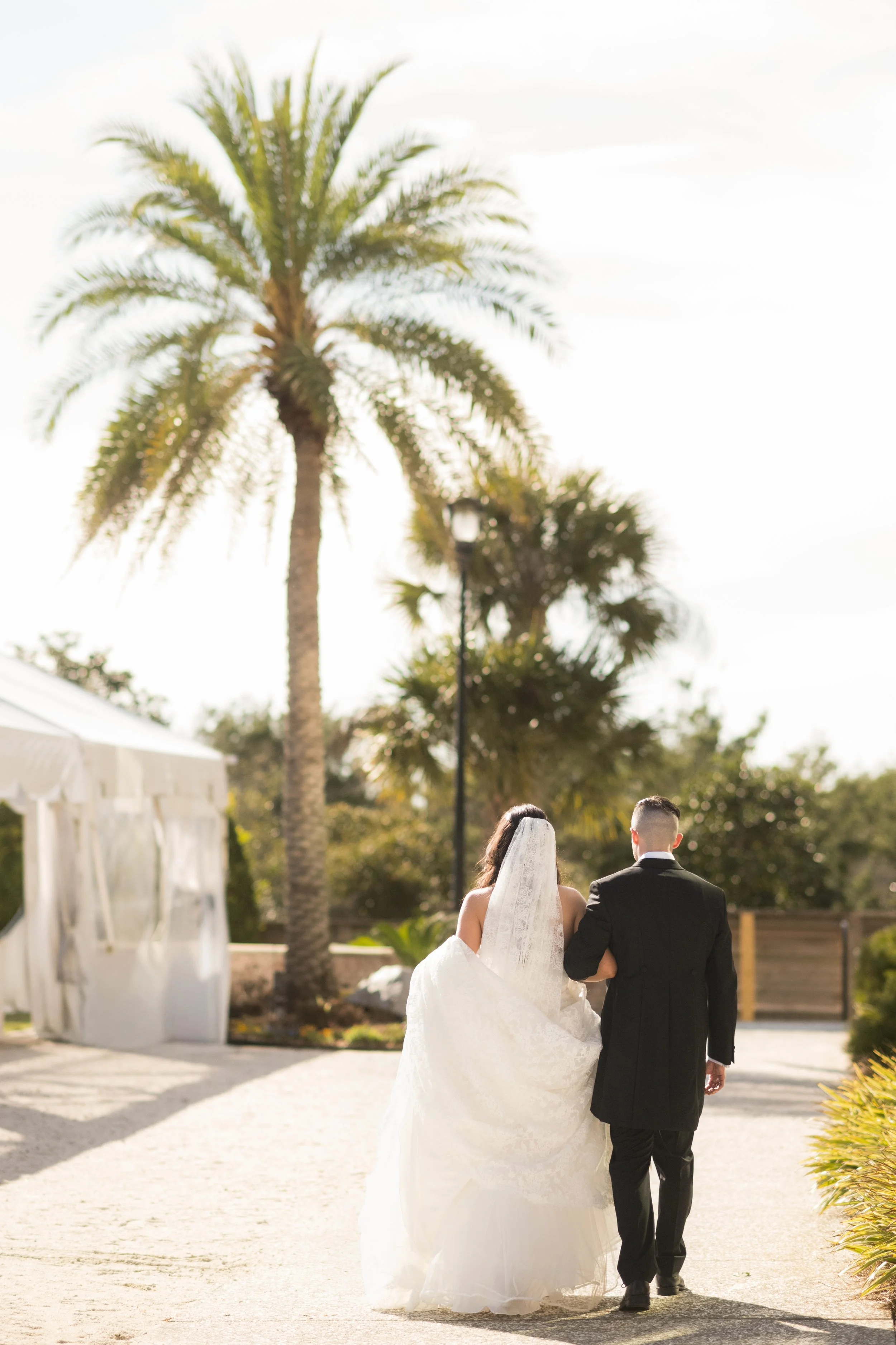 A bride and groom walking arm in arm outdoors during a wedding ceremony, with palm trees and white tents in the background.