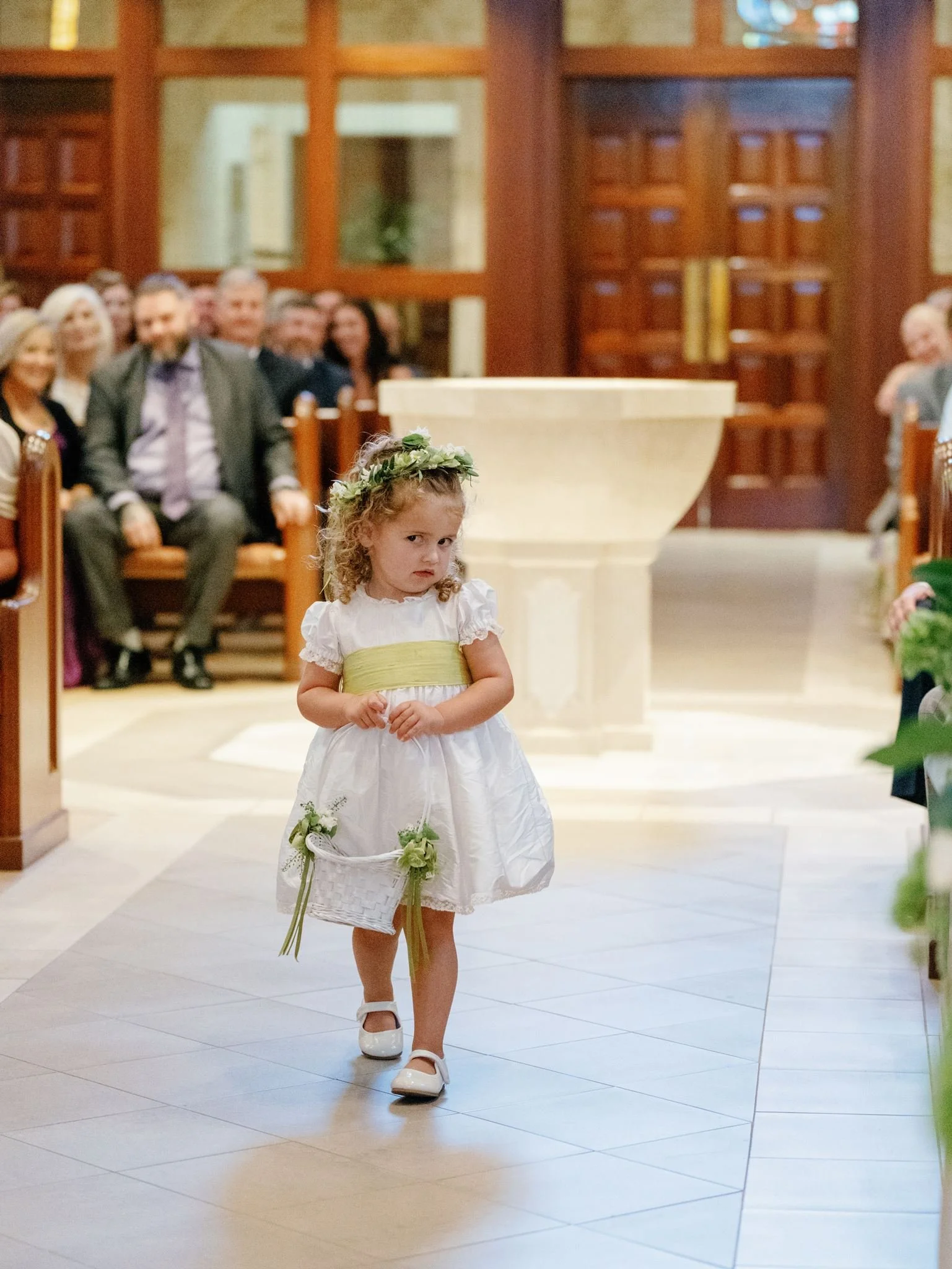 Young girl in white dress with green sash, floral headband, and white shoes walking down aisle in church with attentive audience in background.