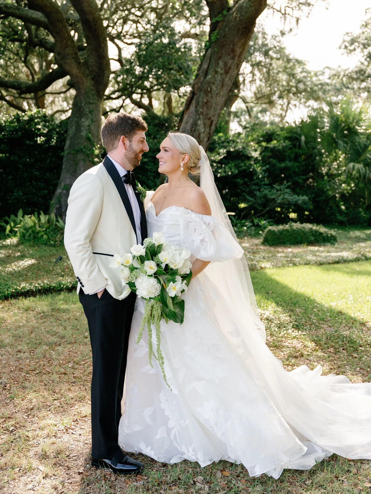 A bride and groom standing outdoors under a large tree, facing each other and smiling, with the bride holding a bouquet of white flowers. The bride is wearing a white off-shoulder wedding gown and veil, while the groom is in a cream tuxedo with black