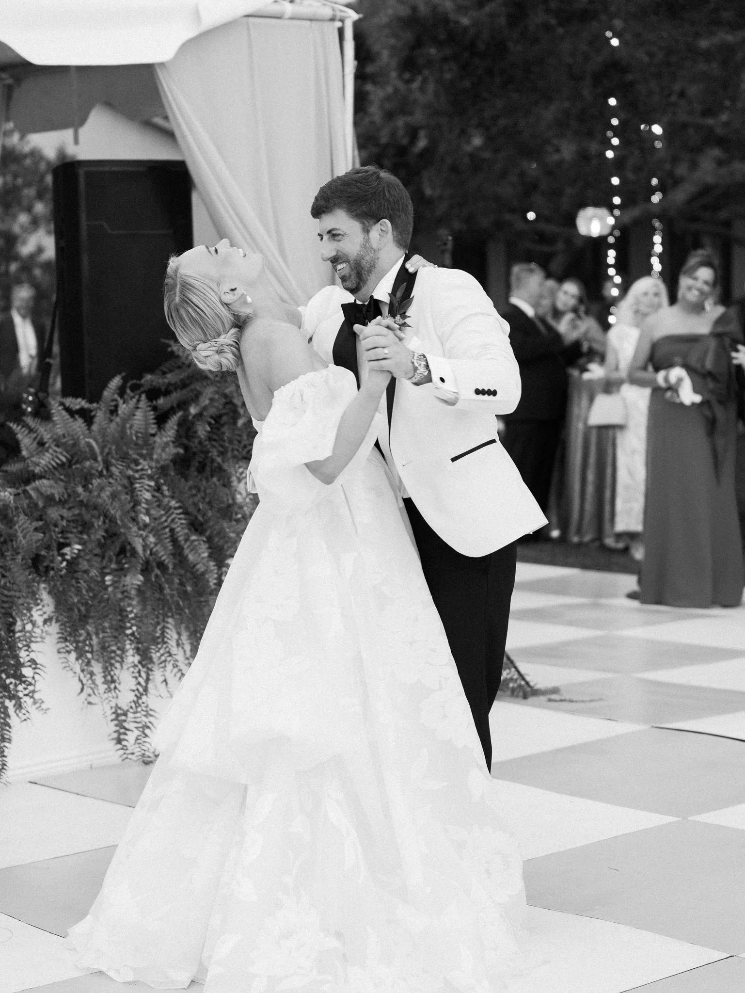 A bride and groom are dancing at their wedding reception. The bride is wearing a strapless wedding gown, and the groom is dressed in a white tuxedo jacket with a black bowtie. They are smiling and looking at each other affectionately. In the backgrou