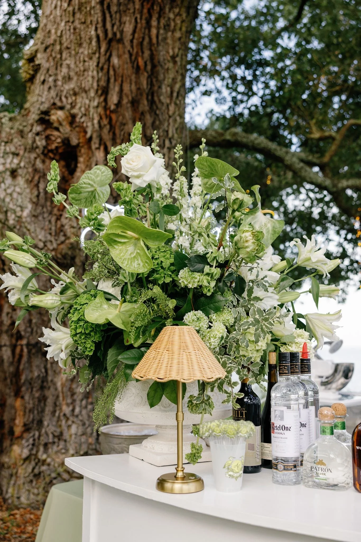 A floral arrangement on a white table, with bottles of alcohol and a small table lamp, outdoors near a large tree with green leaves.