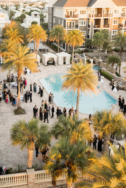 People gathered at an outdoor pool area for a wedding or event, surrounded by palm trees and multi-story buildings.