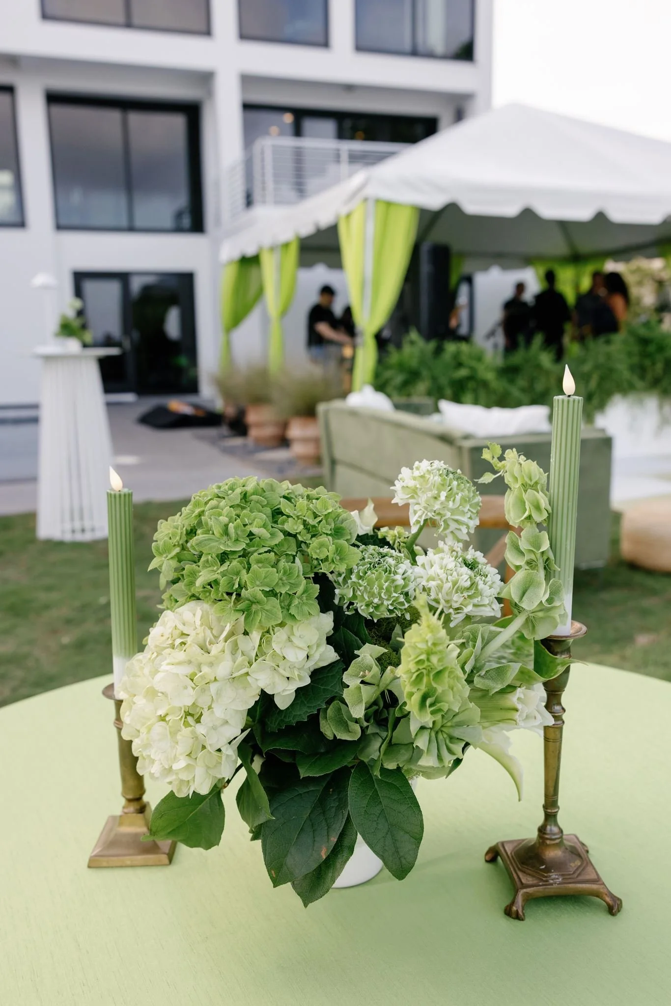 Close-up of a floral centerpiece featuring white and green hydrangeas with large leaves, placed on a light green table. In the background, there is an outdoor setting with a white canopy tent decorated with green drapes, and blurred silhouettes of pe