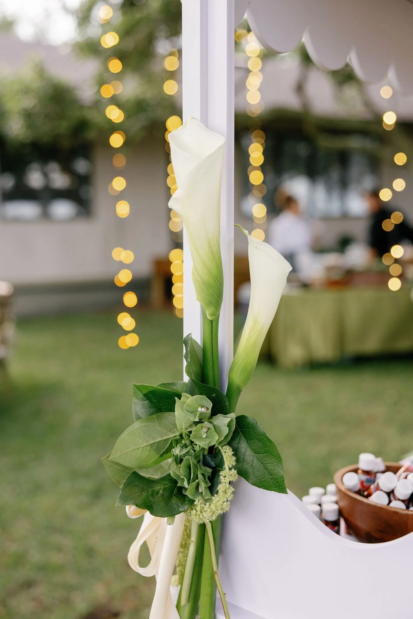 Close-up of a white flower with green leaves, decorated with ribbons and surrounded by small white pills in a bowl, with blurred string lights and outdoor setting in the background.