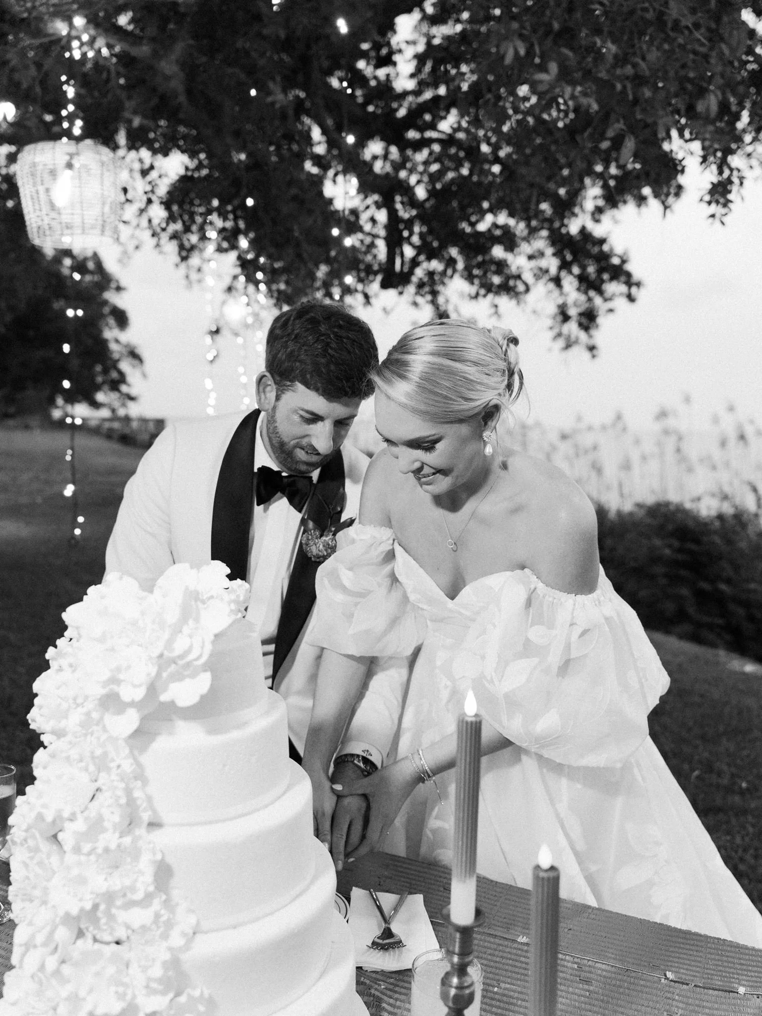 A bride and groom cutting their wedding cake outdoors during the evening, with a decorated tree and string lights in the background.