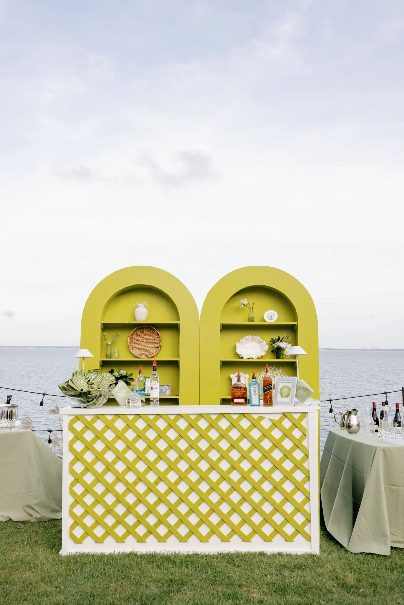Yellow outdoor bar with arched shelving against a body of water under a cloudy sky, decorated with bottles, vases, and decorative objects, with tables and chairs nearby.