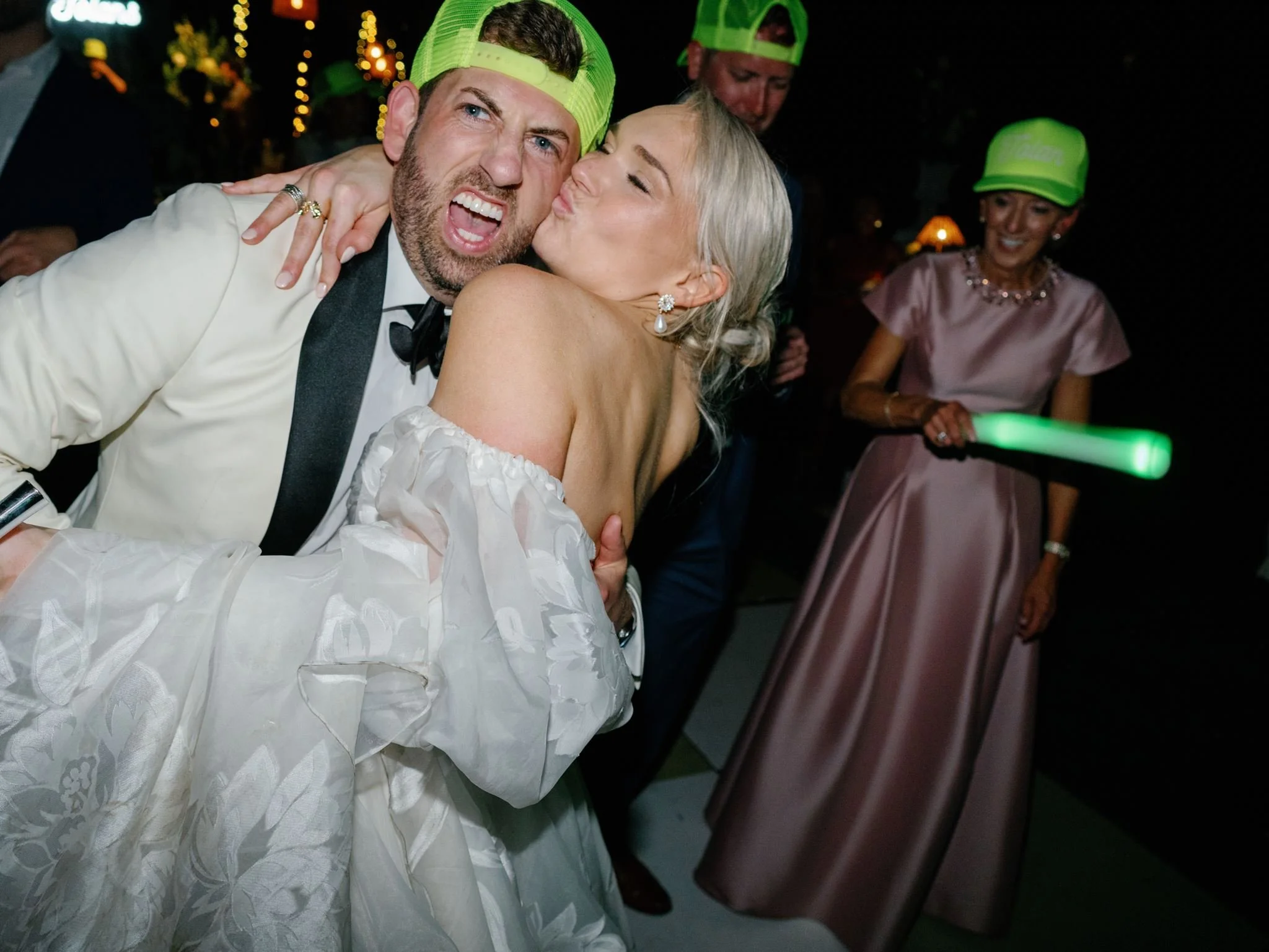 A young man in a tuxedo and a woman in a white dress are hugging and kissing at a celebration or event, with other people in the background wearing bright green caps.