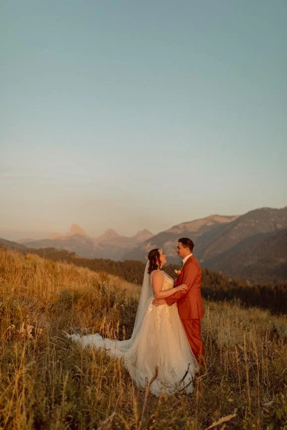 A bride and groom stand facing each other in a grassy field with mountains in the background during sunset.