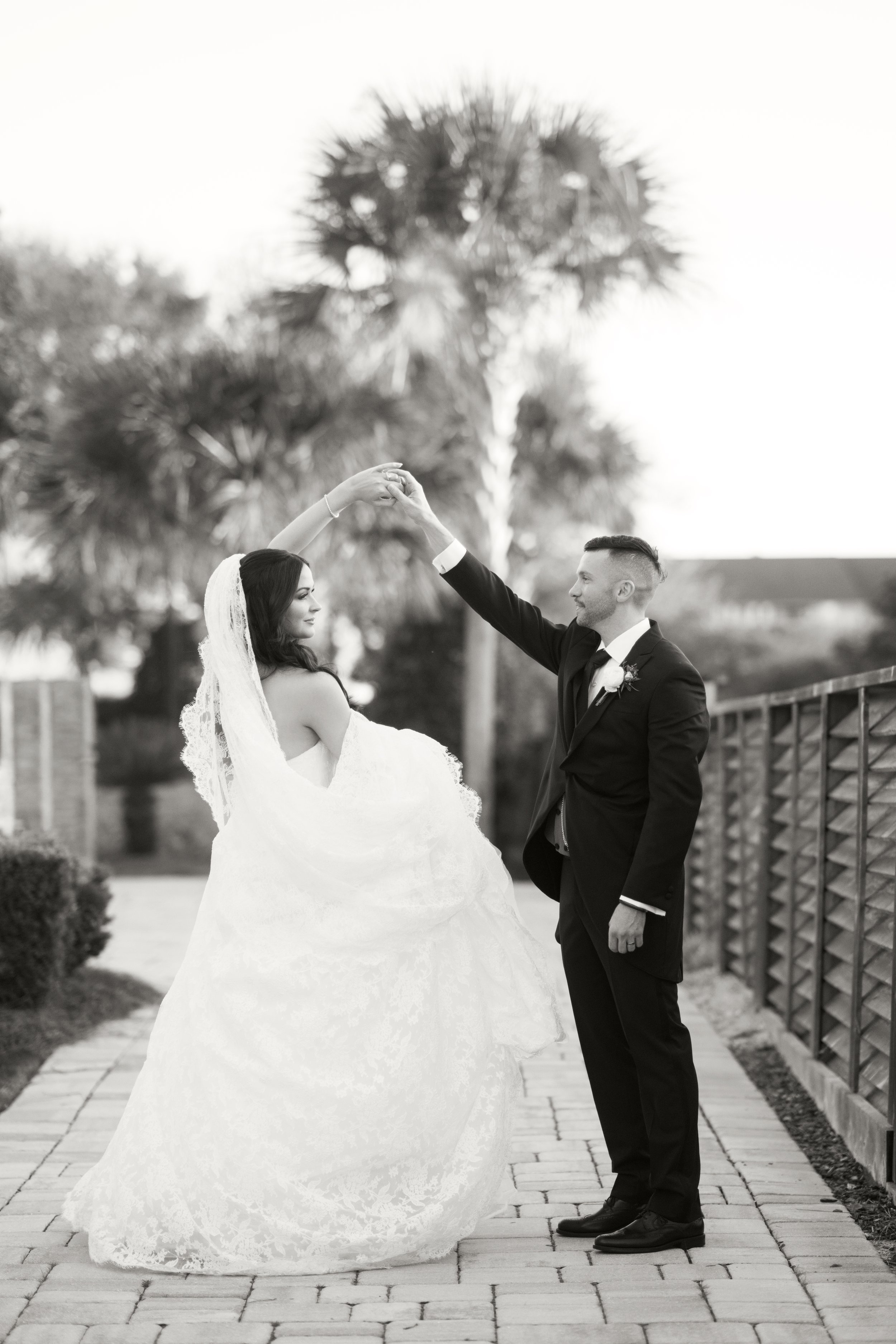 A bride and groom dancing outdoors, with the groom holding the bride's hand as she twirls in her wedding dress, against a background of trees and a wooden fence, in a black-and-white photograph.