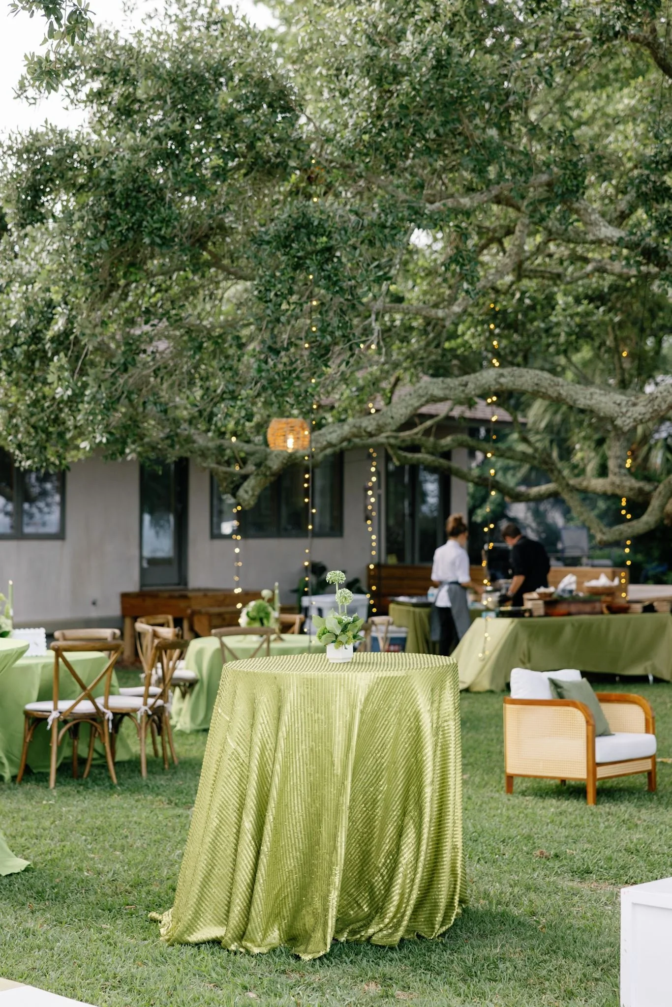 Outdoor garden scene with a table covered in a yellow-green tablecloth and a centerpiece of white flowers, surrounded by chairs, with a large tree decorated with string lights and hanging lanterns, and a house in the background.