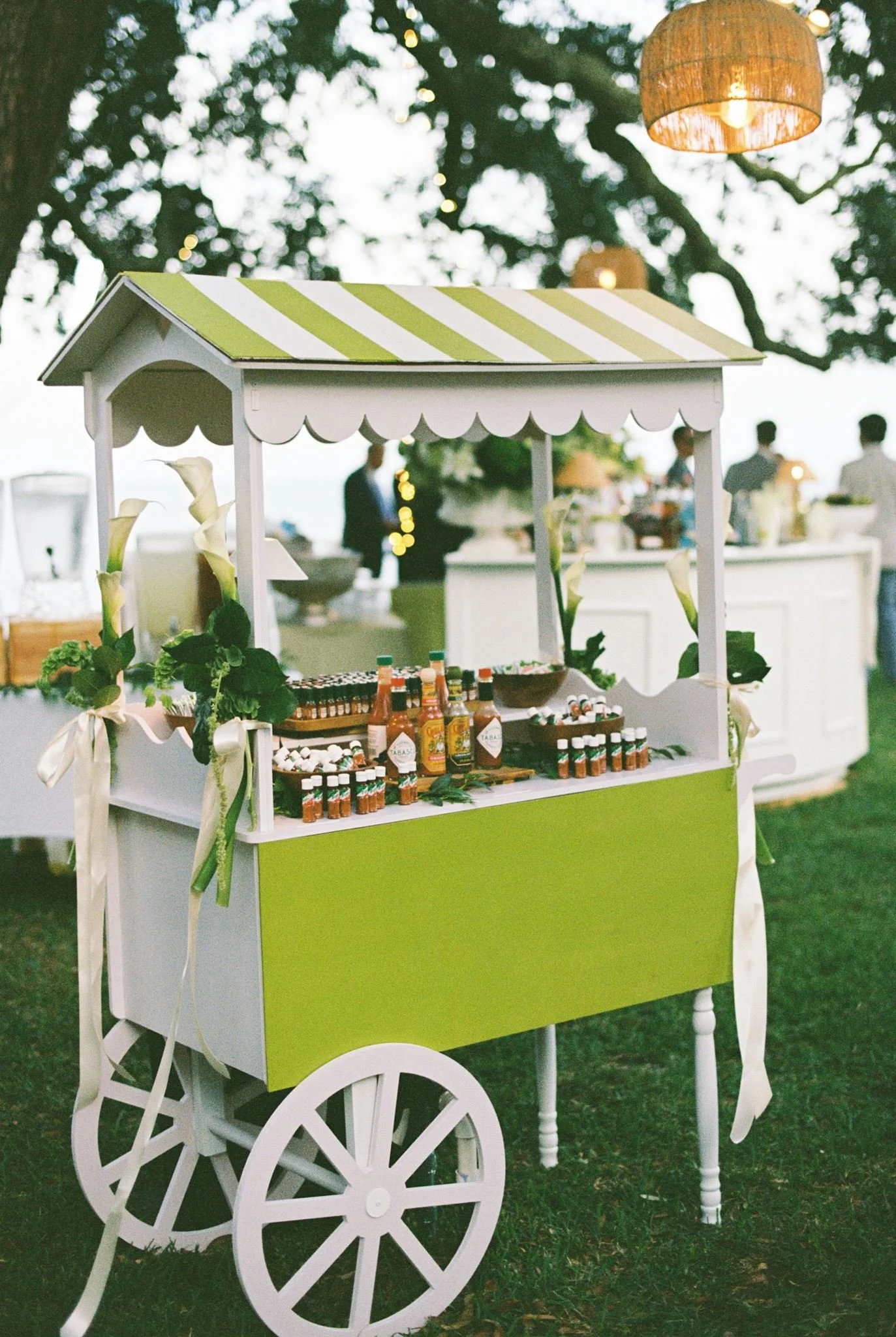 A white and green wooden cart with wheels, decorated with flowers and ribbons, selling hot sauces and condiments at an outdoor event during the daytime. In the background, there are trees and people dressed in formal attire.
