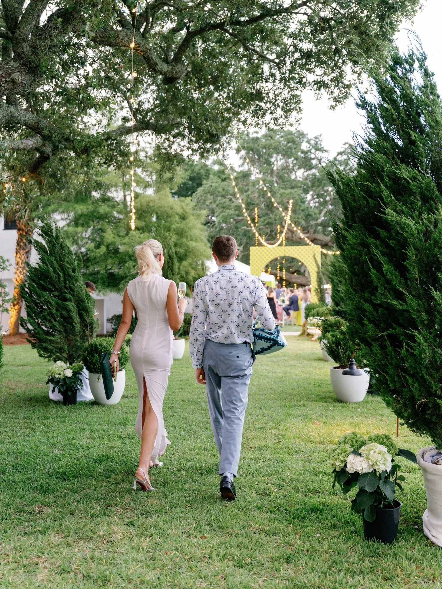 A couple dressed in formal attire walking through a garden at an outdoor event during evening time, with decorative string lights hanging above.