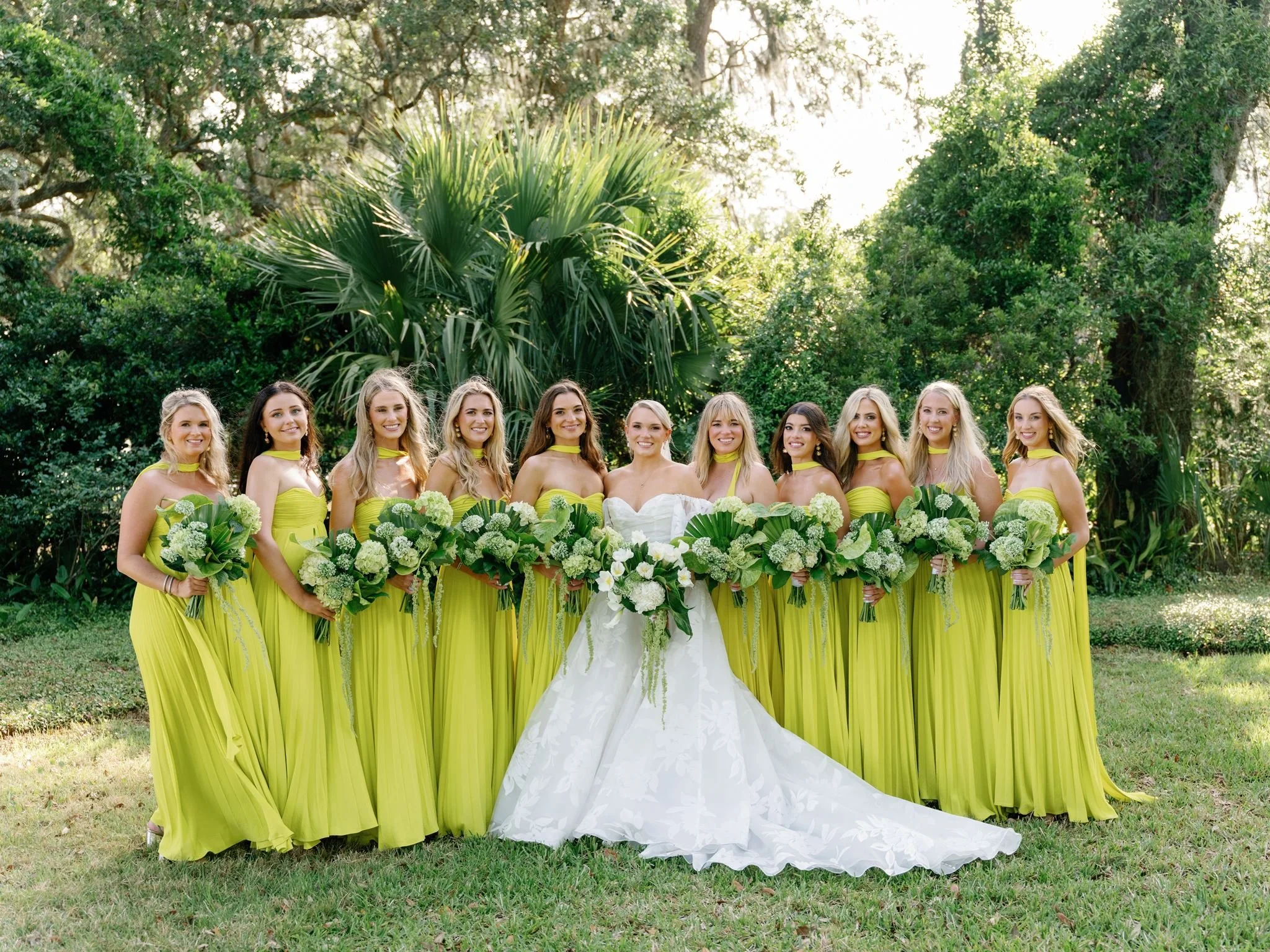 A bride and ten bridesmaids standing outdoors in front of greenery and trees, with sunlight filtering through. The women are dressed in matching bright yellow dresses and are holding bouquets of white flowers and green foliage.