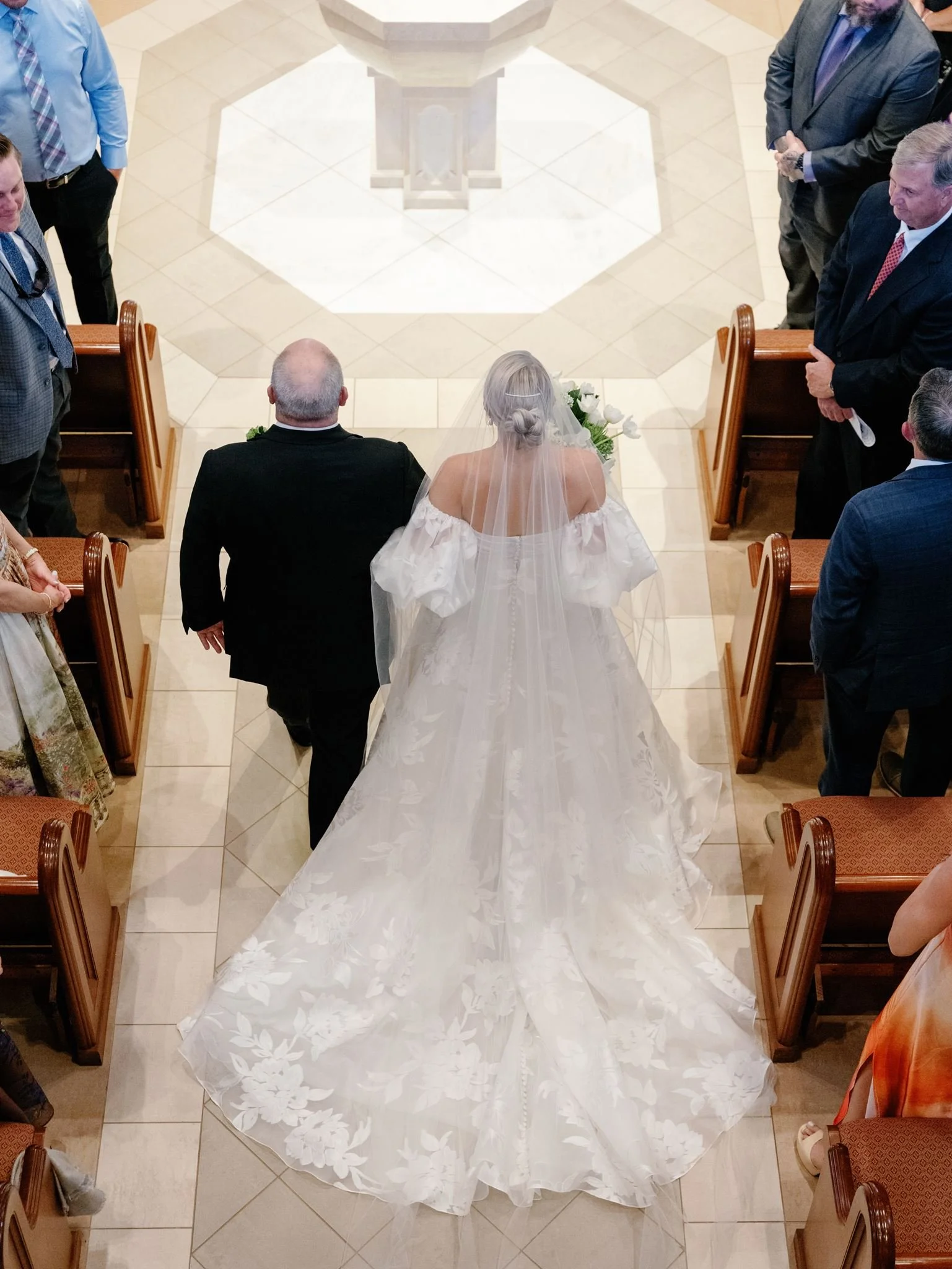 A bride in a white wedding dress with a long train and veil walking down the aisle in a church, accompanied by her father. Guests are standing on either side, watching her walk.