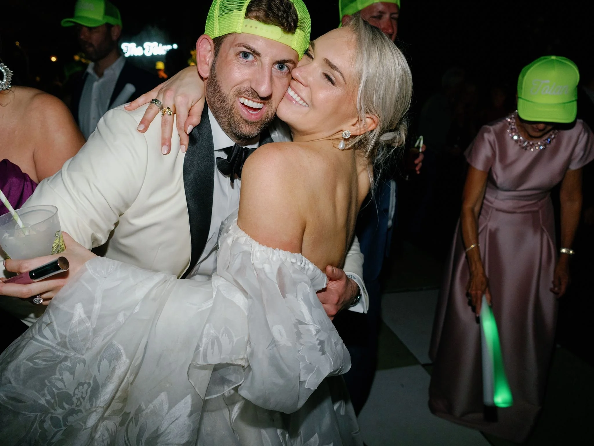 A man and woman hugging and smiling at a party or celebration, with other people in the background wearing neon green hats and dancing.