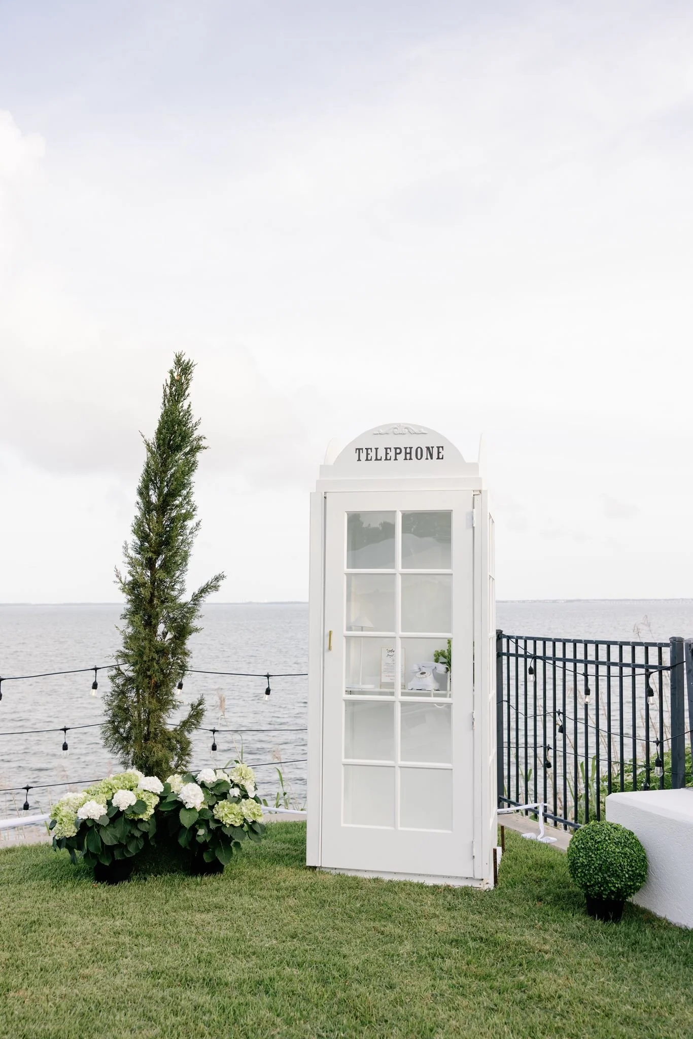 A white telephone booth decorated for a wedding by the water, with a small potted plant inside, standing on a grassy area next to a black metal fence and string lights, with a tree and clouds in the sky in the background.