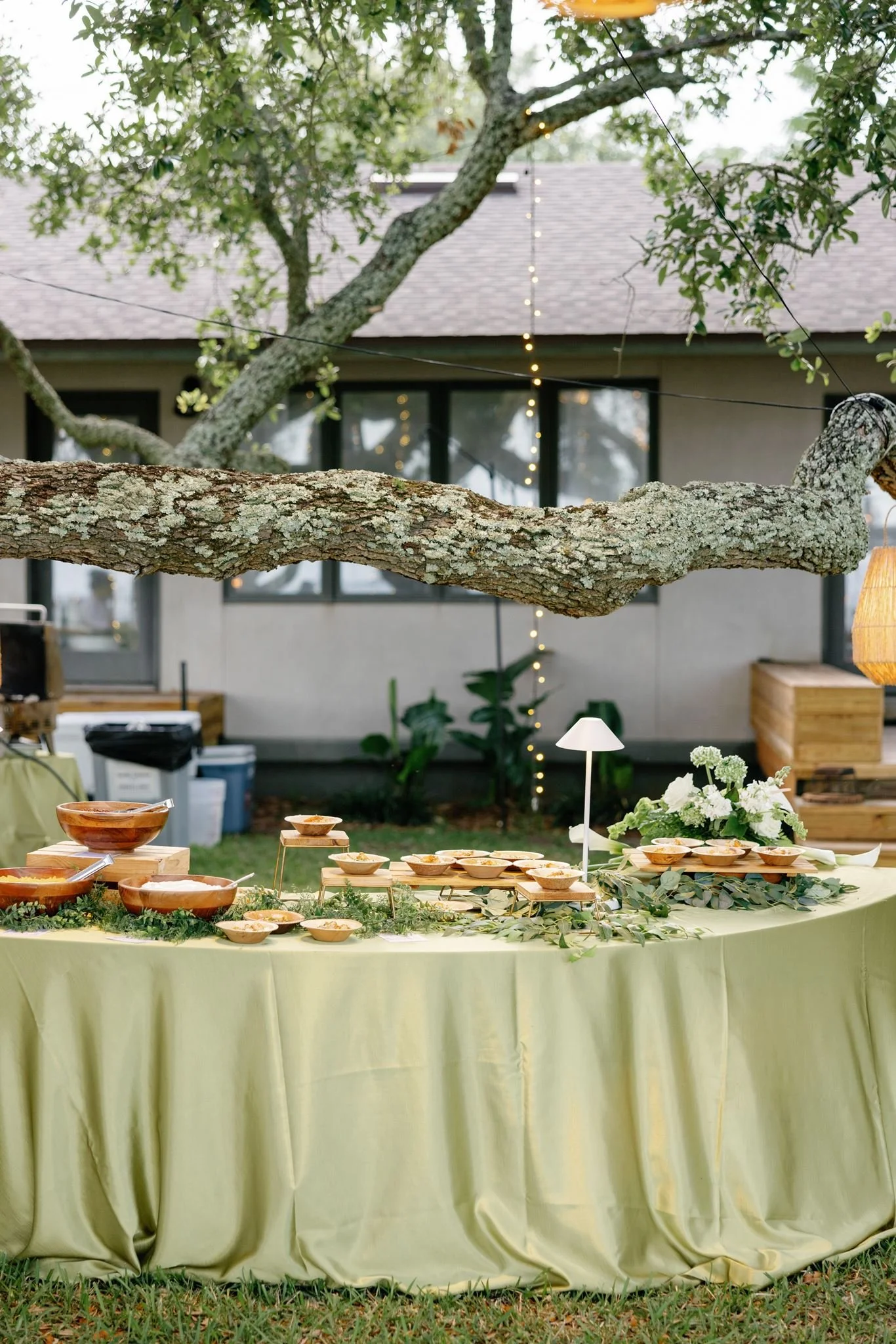 Outdoor food table with bowls of food, surrounded by greenery and trees, with a house in the background.