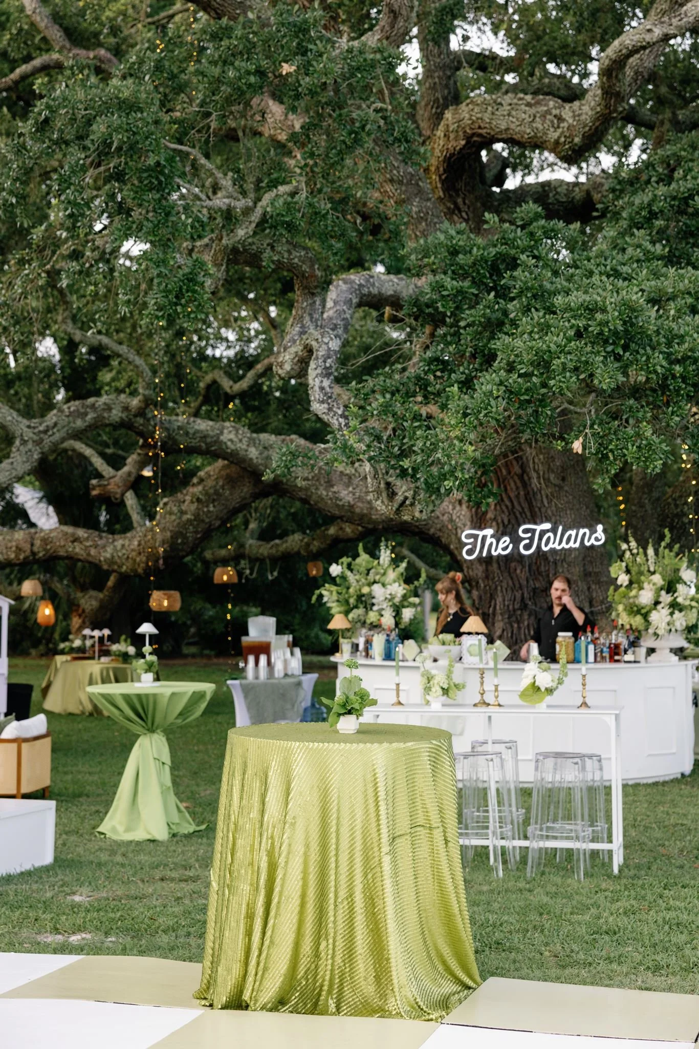 Outdoor event with round tables covered in green and yellow tablecloths, a bar area under a large tree, and string lights decorations.
