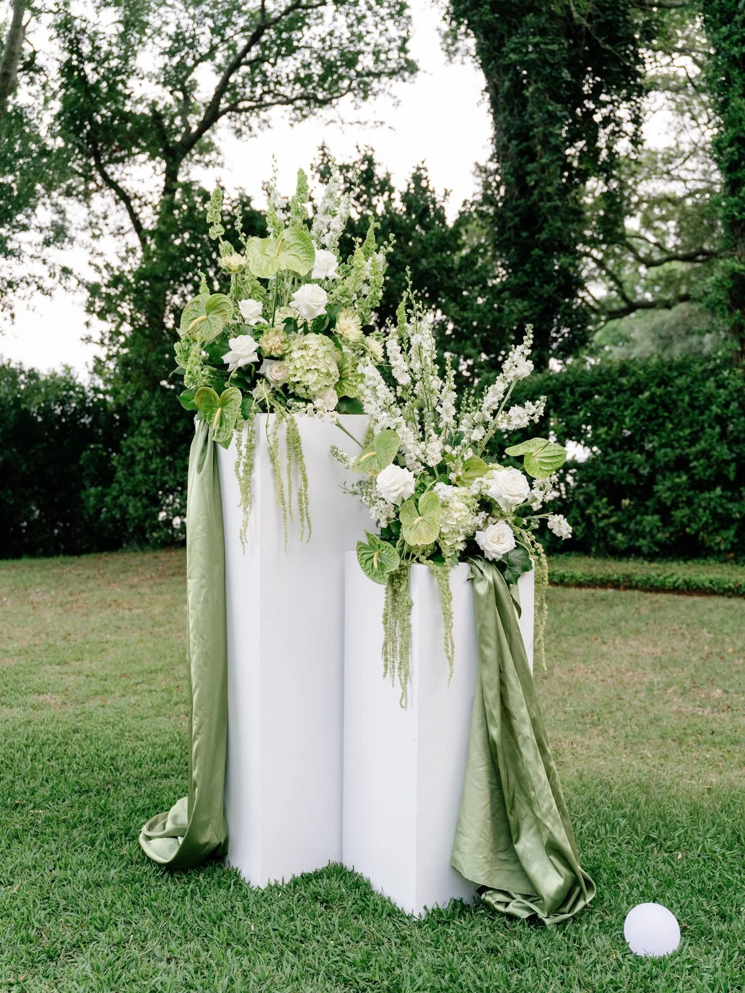 Elegant outdoor wedding altar decorated with white and green flowers, set on green grass with trees in the background and a white ball on the ground.