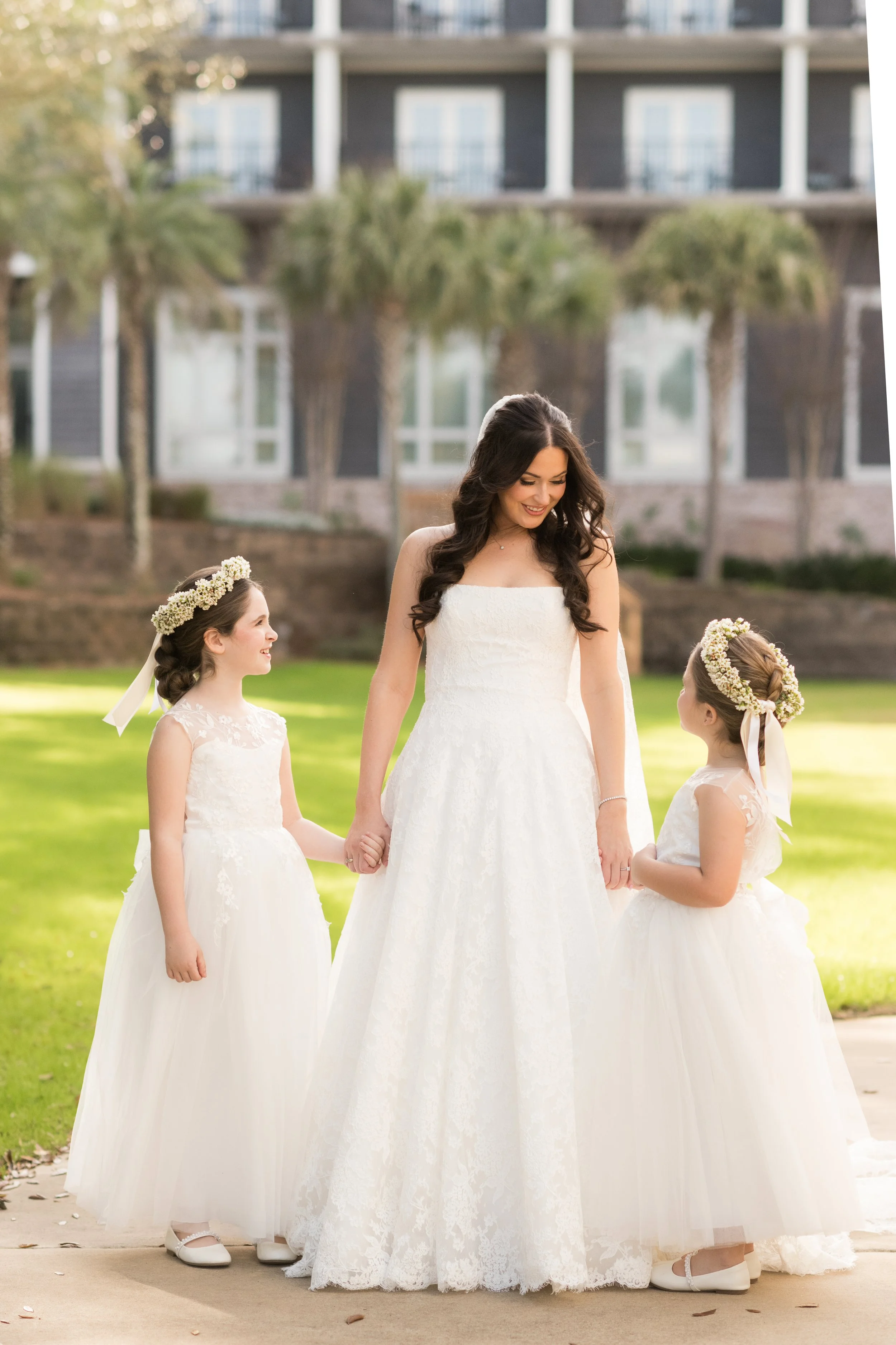 A woman in a wedding dress holding hands with two young girls in white dresses with floral crowns outdoors with a building in the background.