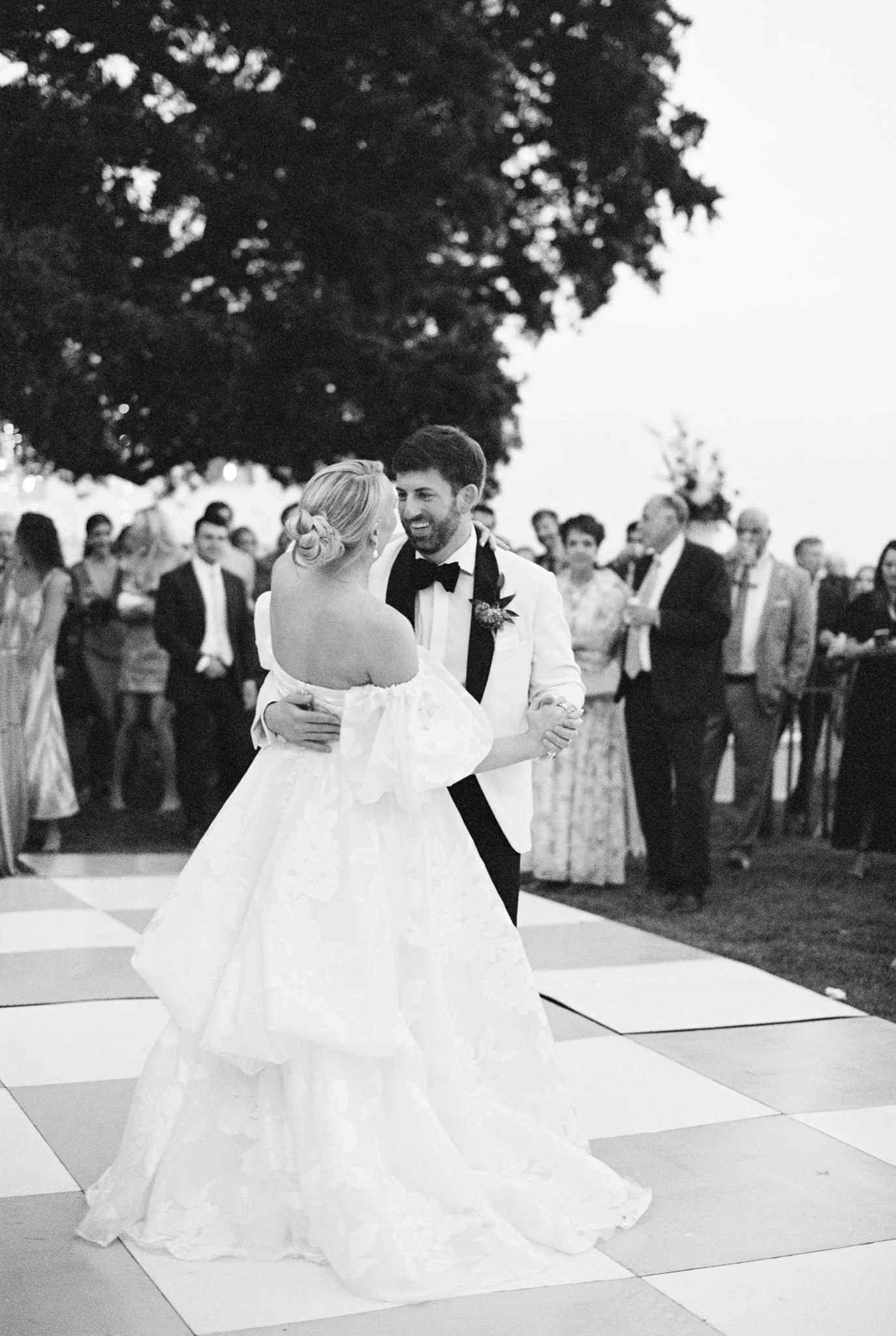 A black and white photo of a bride and groom dancing at their wedding reception outdoors, surrounded by guests. The bride is wearing a strapless gown, and the groom is in a tuxedo. They are smiling at each other while dancing on a checkered dance flo