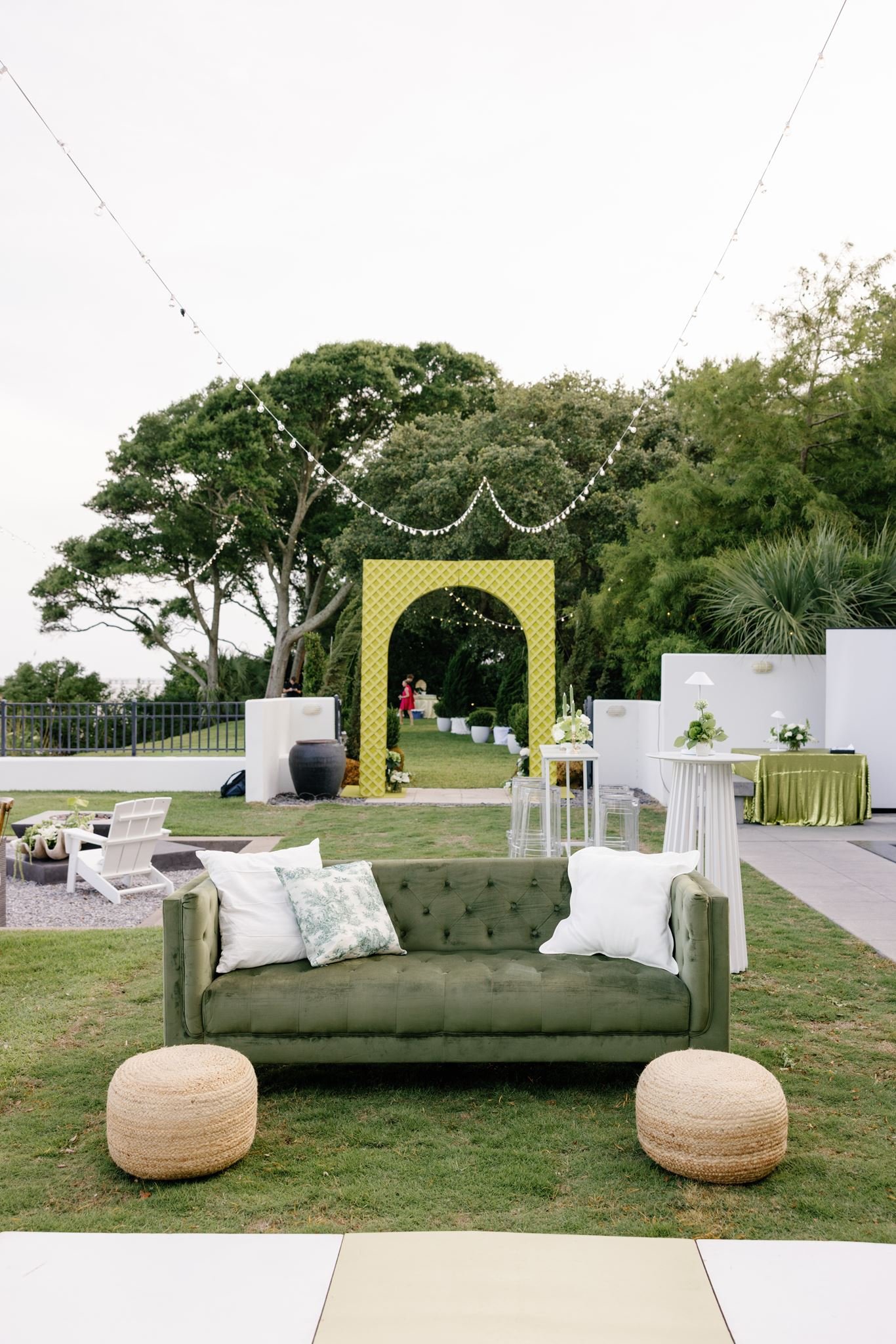 Outdoor event space with a green velvet sofa and two woven poufs in the foreground, a yellow lattice archway in the background, string lights hanging overhead, and decorated tables with green and white accents among lush green trees.