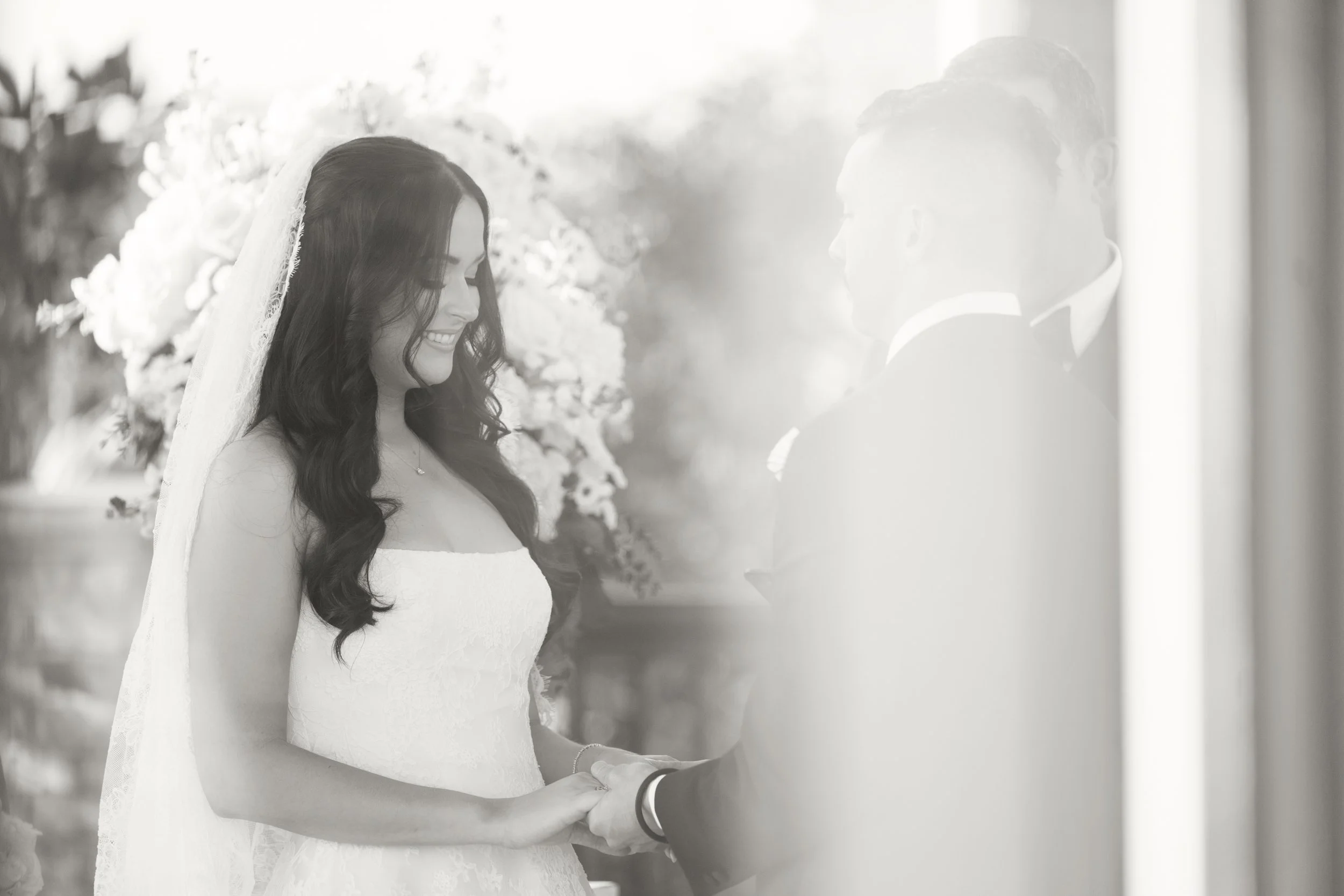 Black-and-white photo of a bride smiling and holding hands with someone during a wedding ceremony, with flowers in the background.