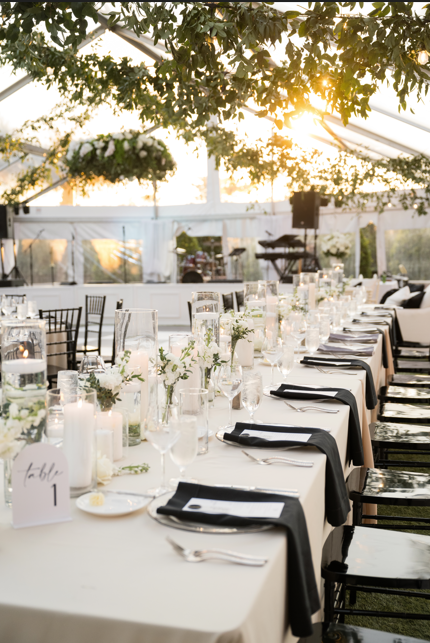 Elegant banquet table set with white tablecloths, black napkins, glassware, candles, and floral centerpieces inside a decorated tent, with sunlight filtering through trees.
