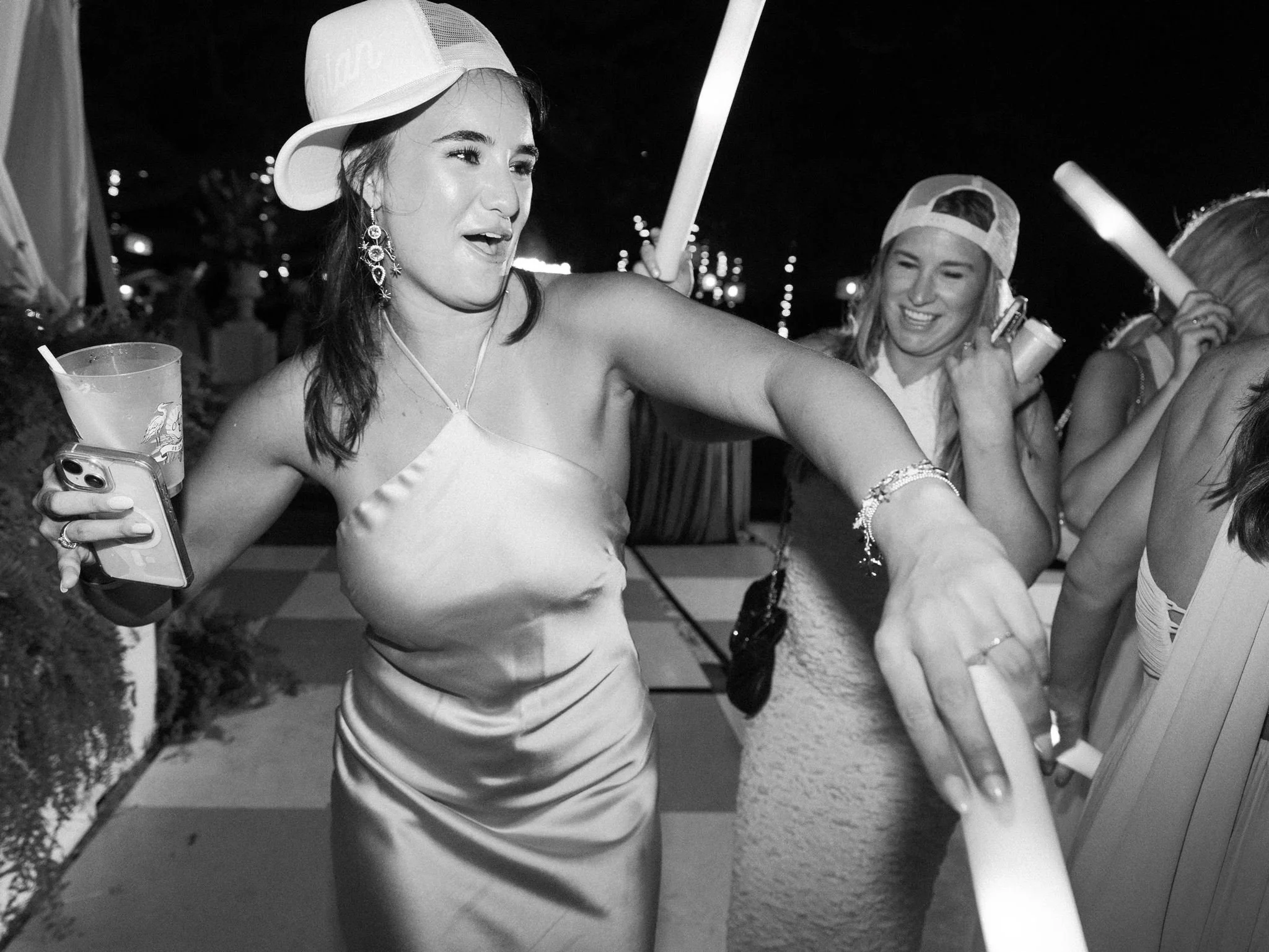 Women celebrating at a party, smiling and holding drinks and glow sticks, with city lights in the background.