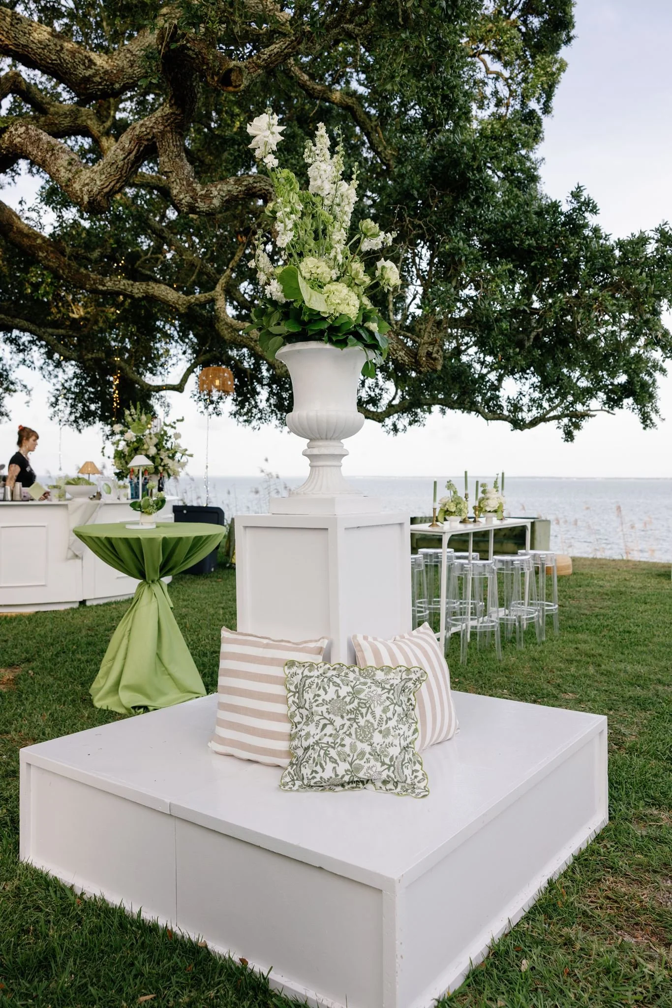 Outdoor event setup with a large white floral arrangement on a tall pedestal, decorated tables, pillows, and a view of the ocean and sky in the background.