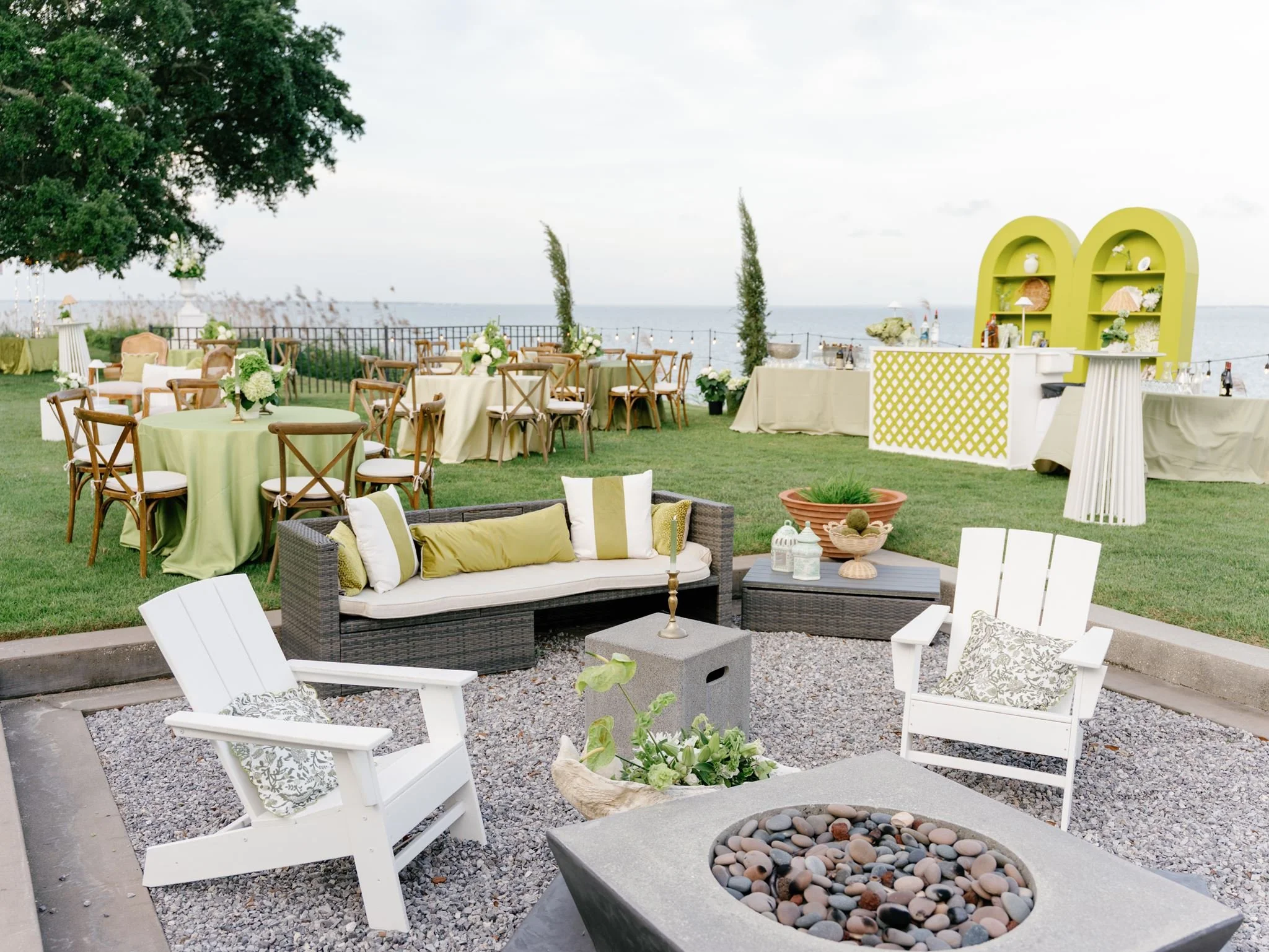 Outdoor wedding reception setup with green and white decor, wooden chairs, tables with floral arrangements, a bar area with shelves and bottles, and a view of the ocean in the background.
