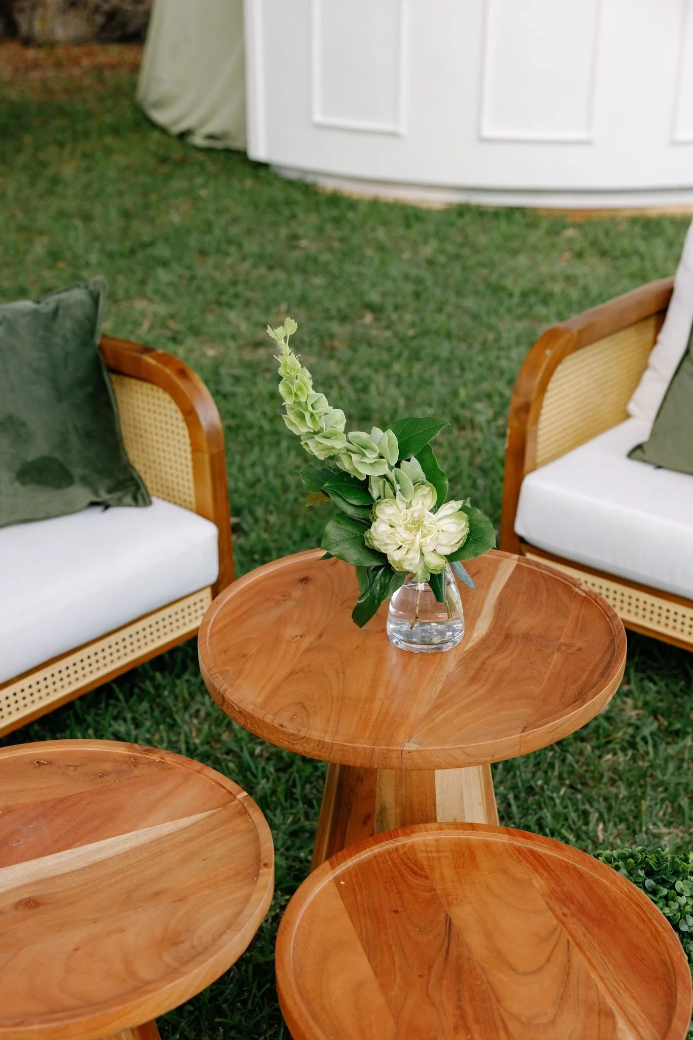 A small glass vase with white flowers and green leaves placed on a round wooden table in an outdoor setting with chairs and grass.