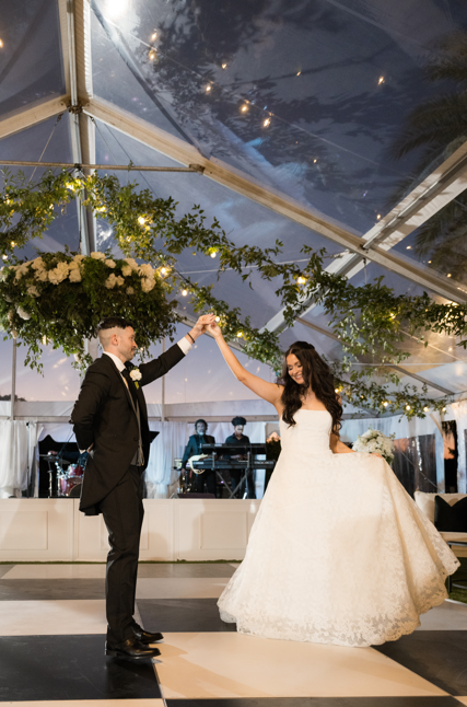 Bride and groom dancing at their wedding reception under a decorated tent with string lights and greenery.