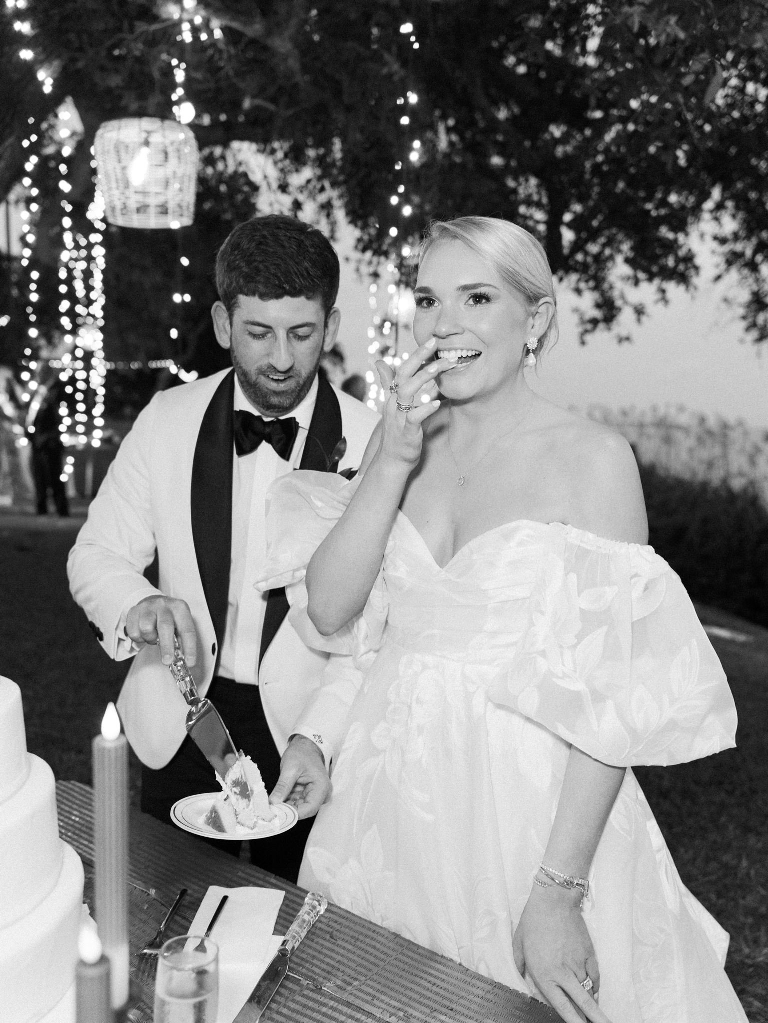A bride and groom at their wedding reception, with the groom cutting the wedding cake. The bride is smiling and wearing a strapless wedding dress, while the groom is in a tuxedo. There are string lights and a hanging lantern in the background.