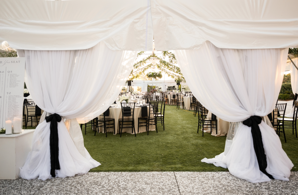 Wedding reception area with round tables and black chairs underneath a decorated white tent with black ties.