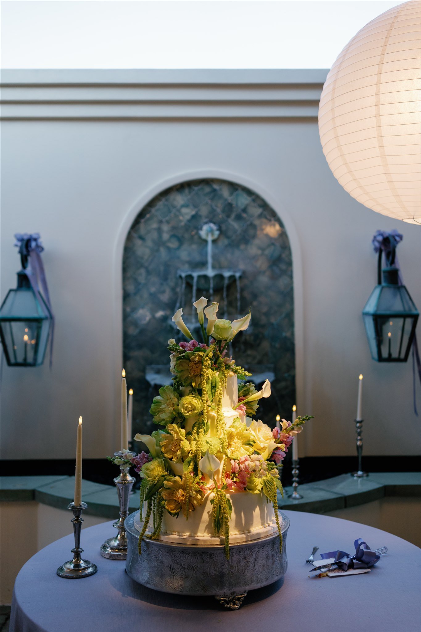 A wedding cake with white frosting and adorned with an elaborate floral arrangement of pink, white, and green flowers. The cake is placed on a round table with silver candelabras holding lit candles around it, in a decorated venue with a fountain on the wall behind and white lanterns on either side.