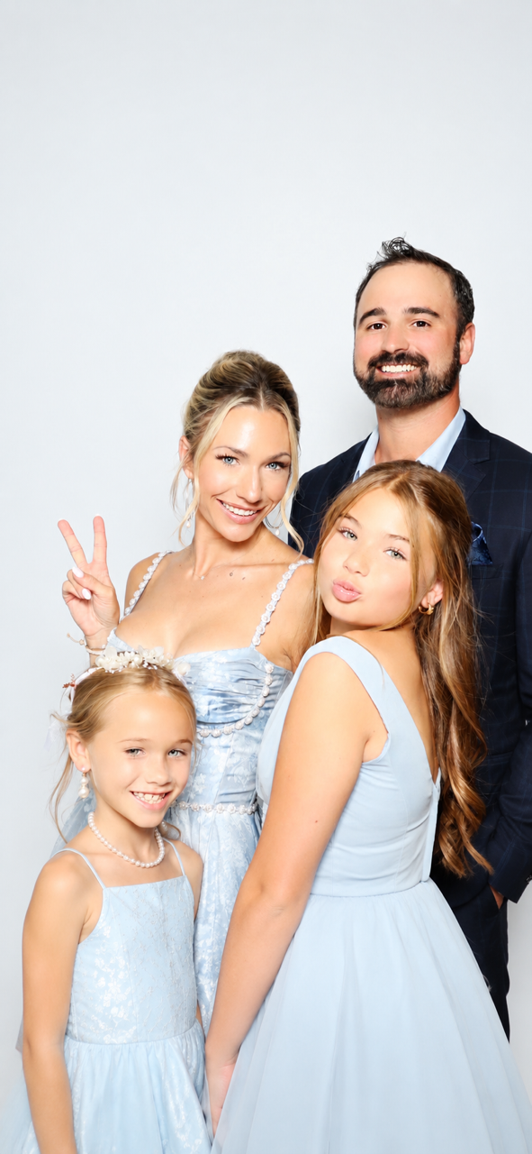 A family of four posing together, with the two young girls wearing light blue dresses and the parents dressed in formal attire, all smiling.