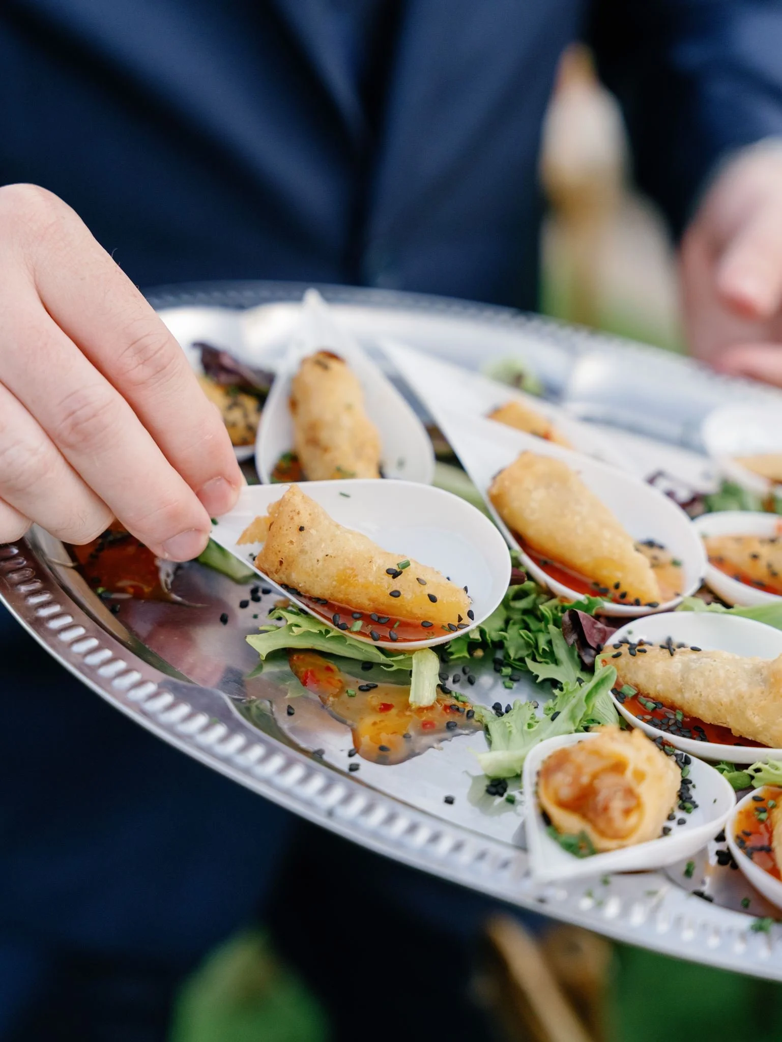 Close-up of a person holding a spoon with fried food on a silver platter with similar fried foods and salad in small bowls.