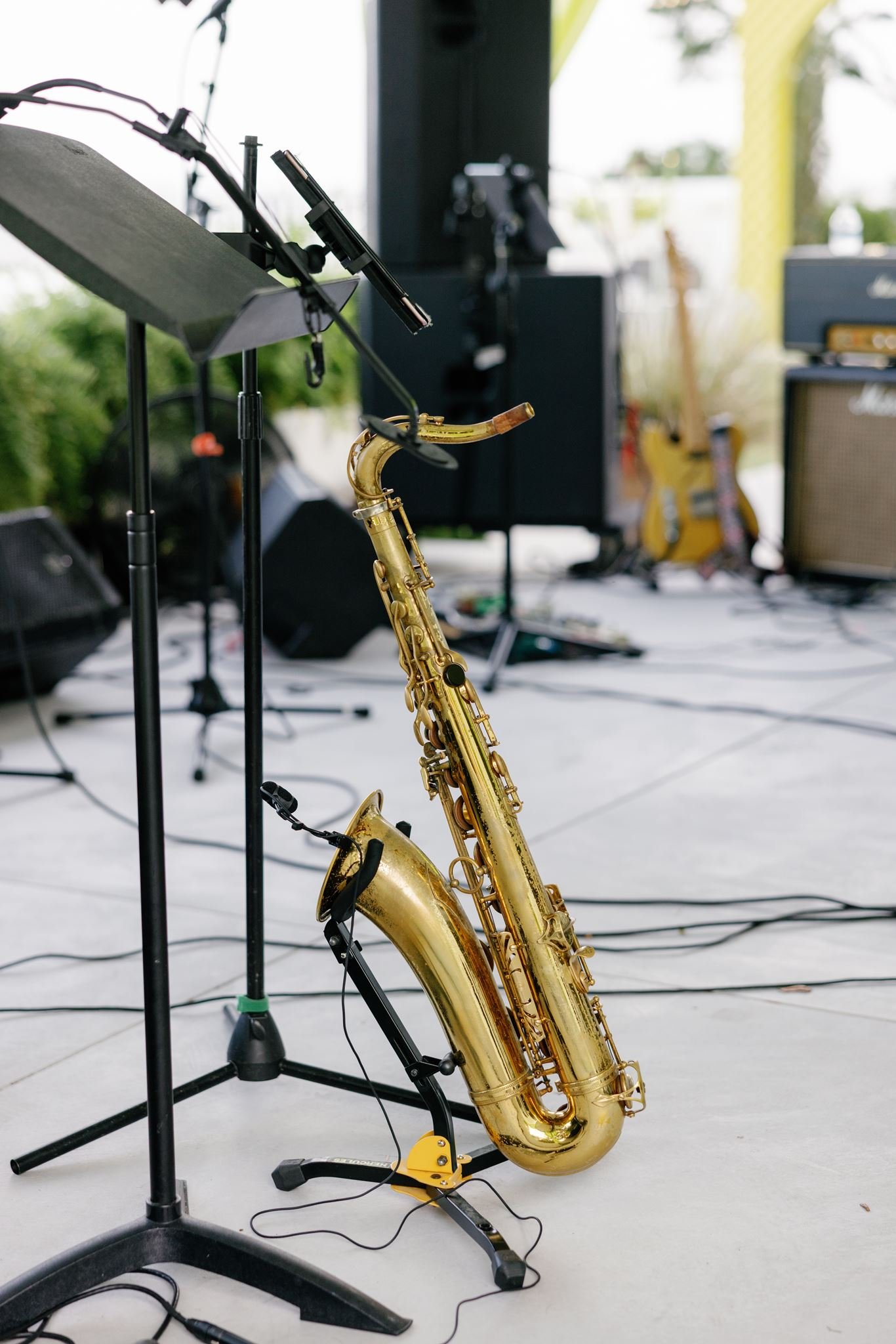 Close-up of a gold saxophone on a stand in front of a music stand, with other musical equipment and instruments in a bright, outdoor setting.